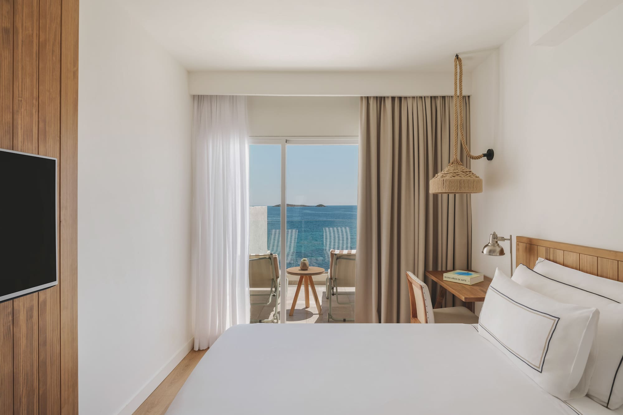 Bright hotel bedroom with sea view balcony, white bed, and wooden decor.
