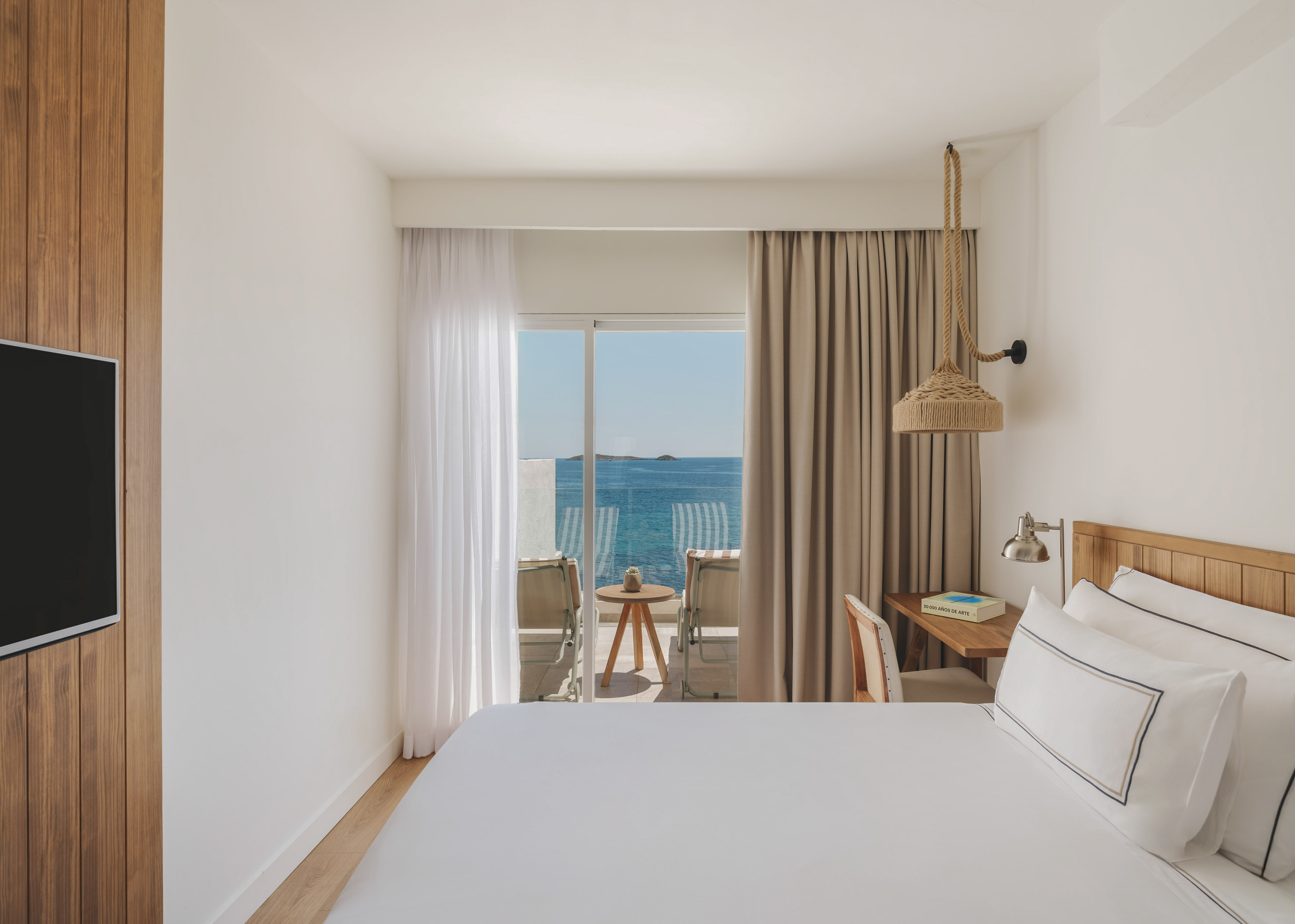 Bright hotel bedroom with sea view balcony, white bed, and wooden decor.