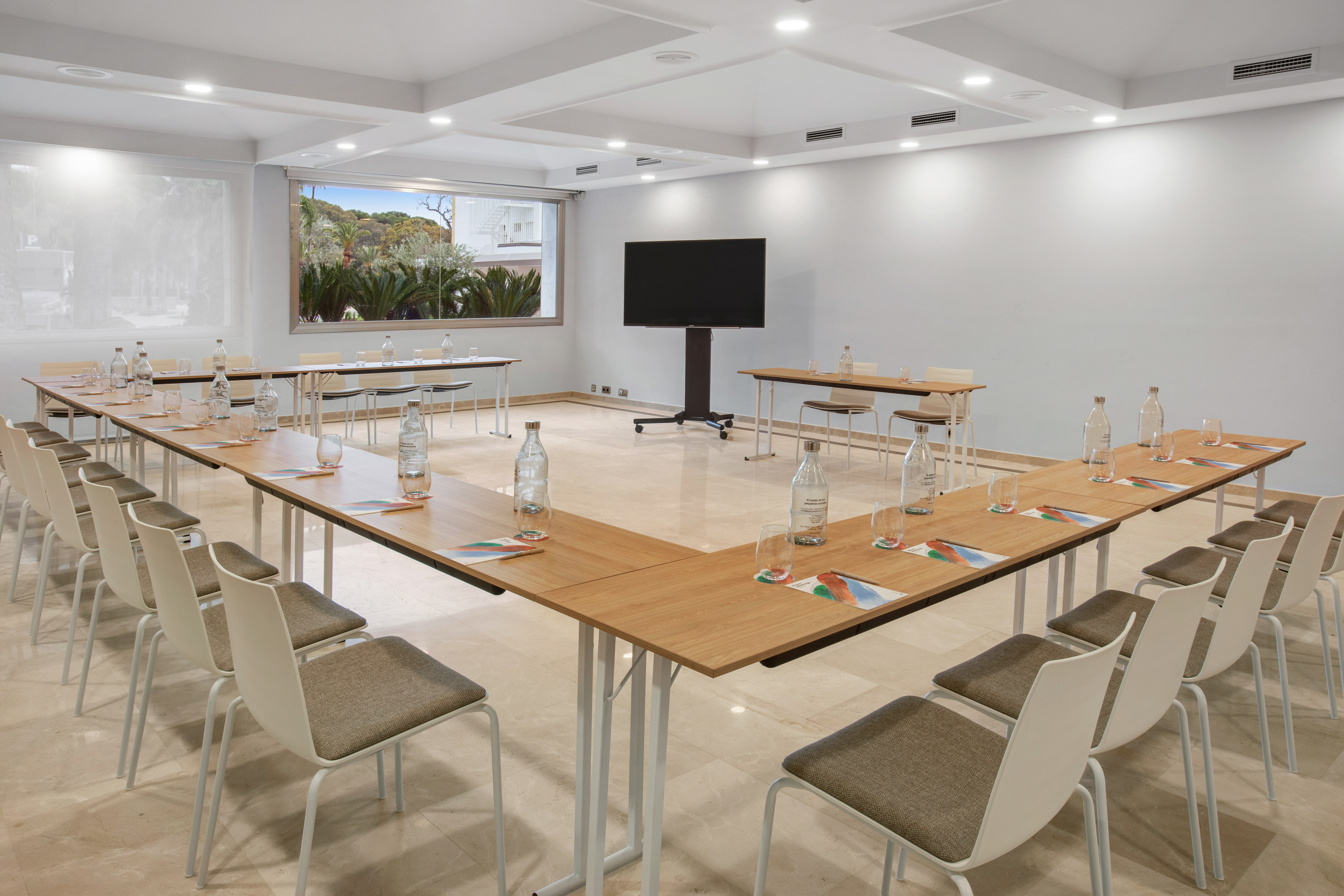 Modern hotel meeting room with U-shaped tables, screen, and palm trees view.