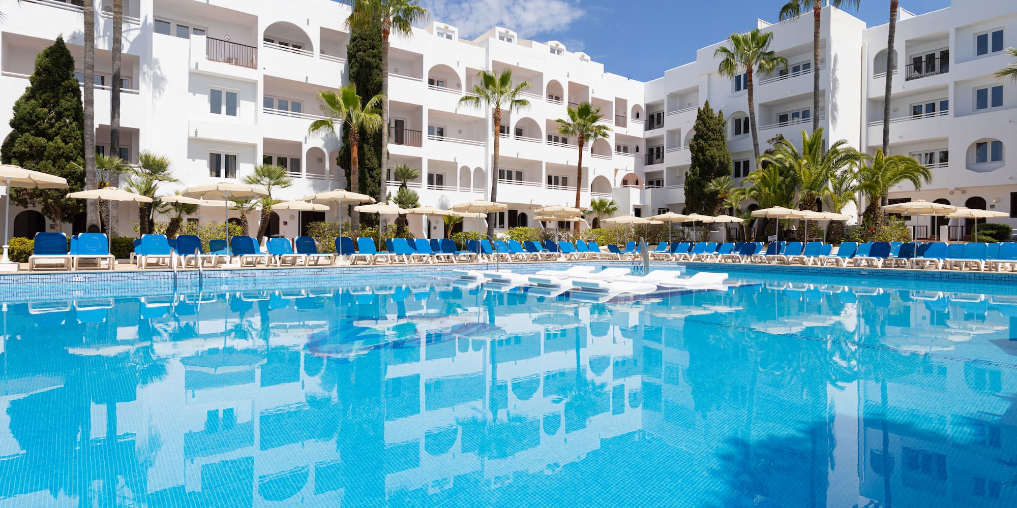 Resort pool, white hotel building, blue sun loungers, and palm trees.
