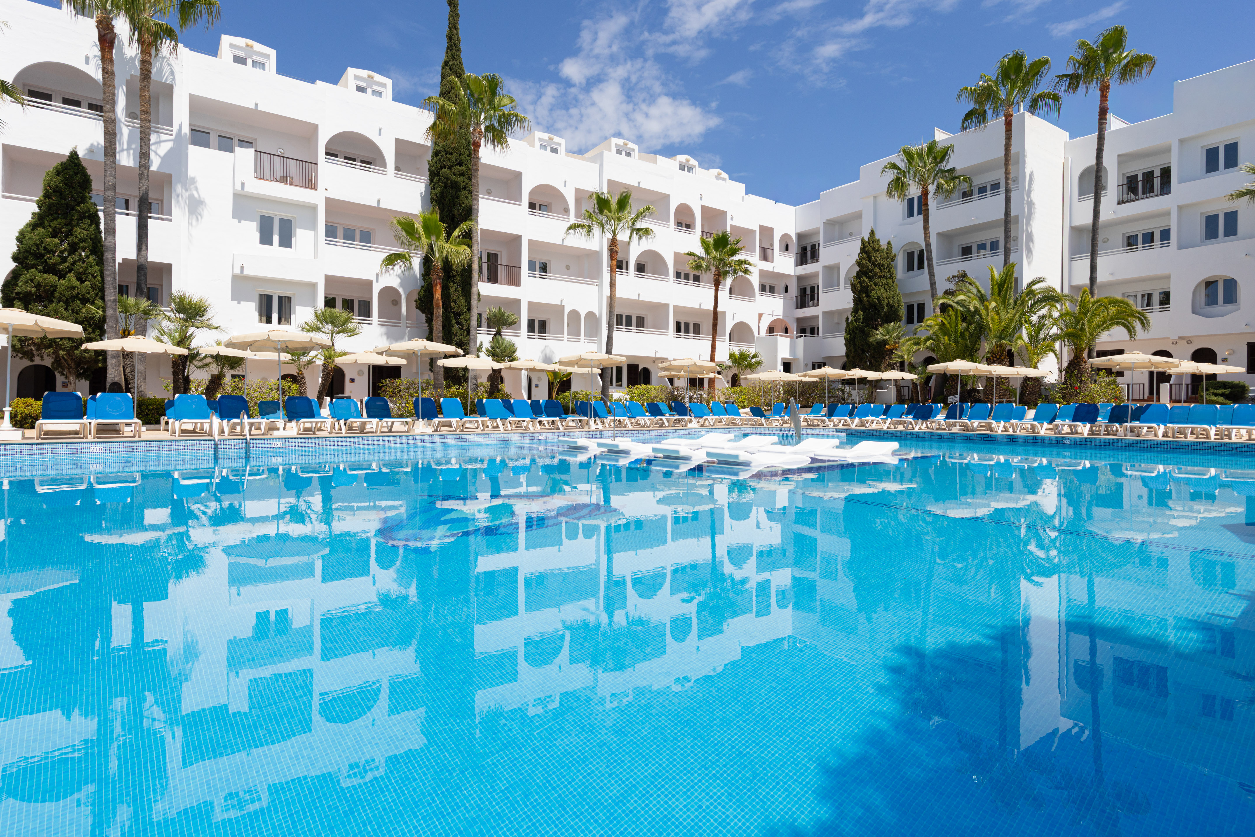 Resort pool, white hotel building, blue sun loungers, and palm trees.