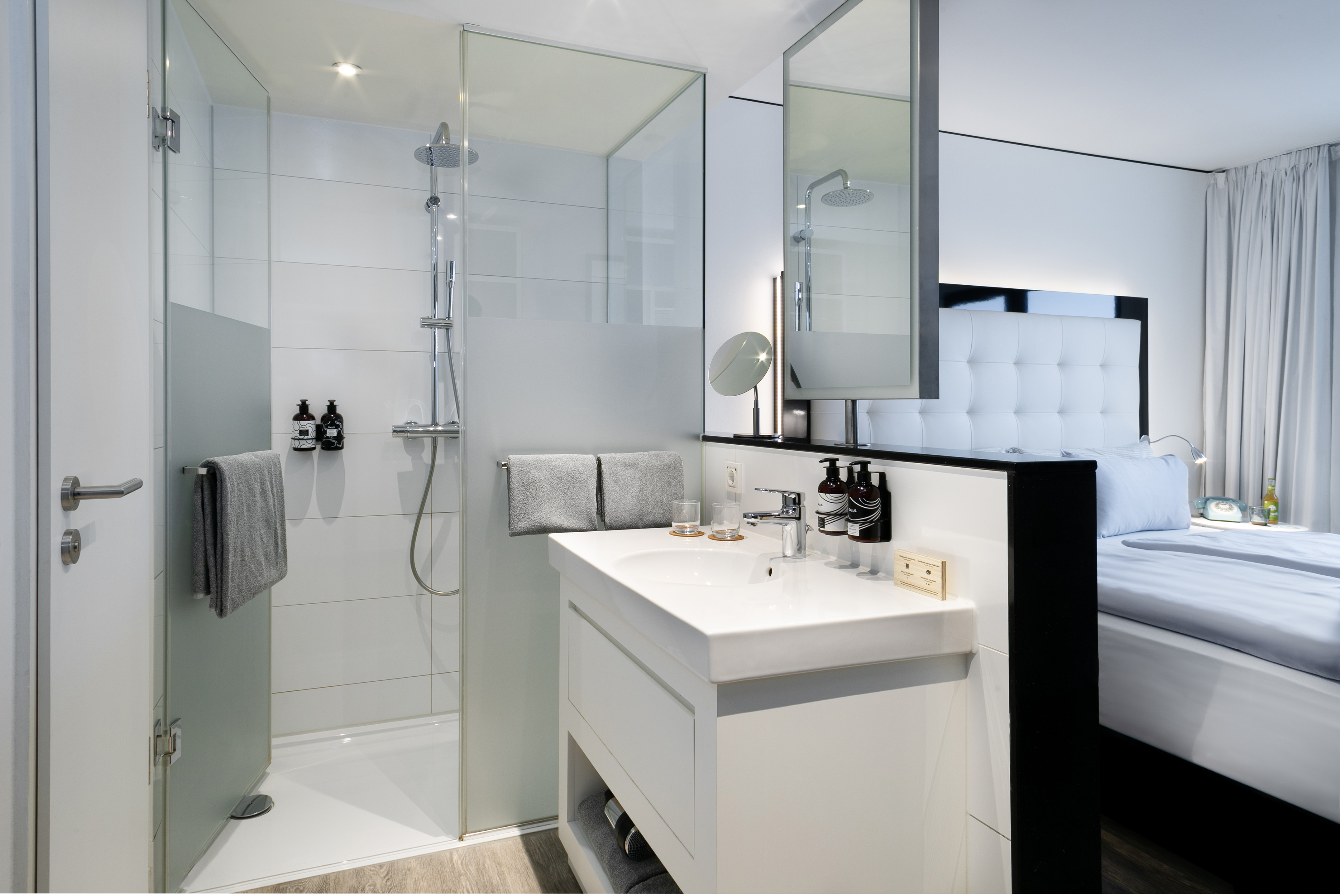 Sleek hotel bathroom with glass shower, white vanity, and modern bedroom visible.