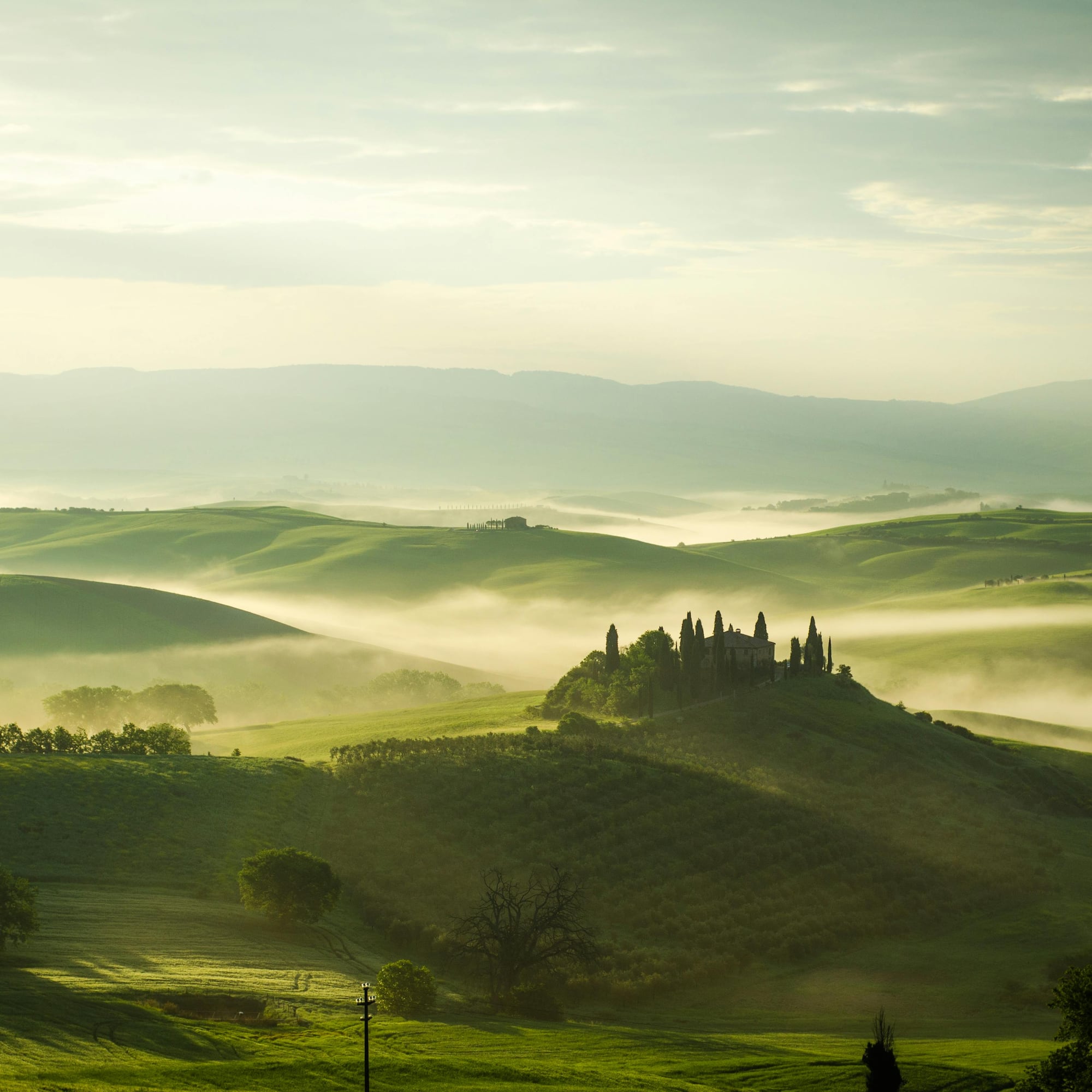 Sunrise over foggy Tuscan hills, with a historic villa and cypress trees.