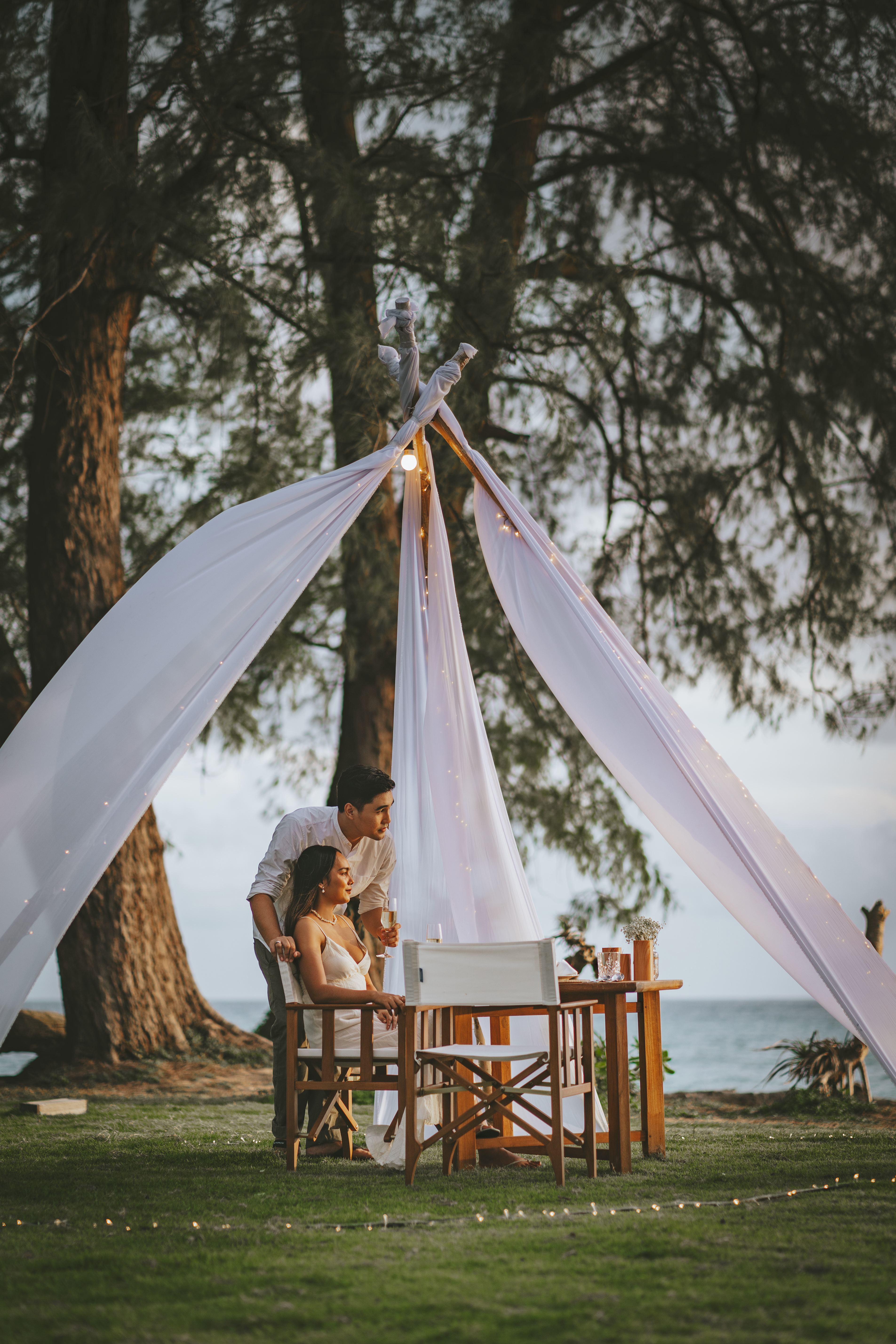 Couple at romantic beach dinner with white drapes, fairy lights, and ocean view.