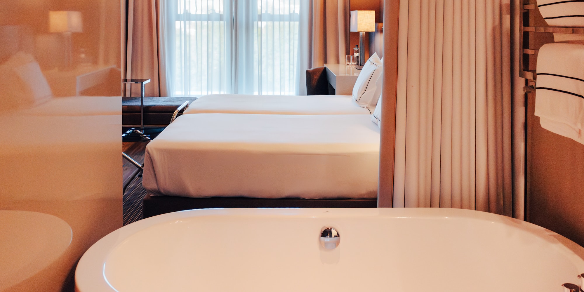 View from white bathtub into a luxury hotel room with twin beds and window.