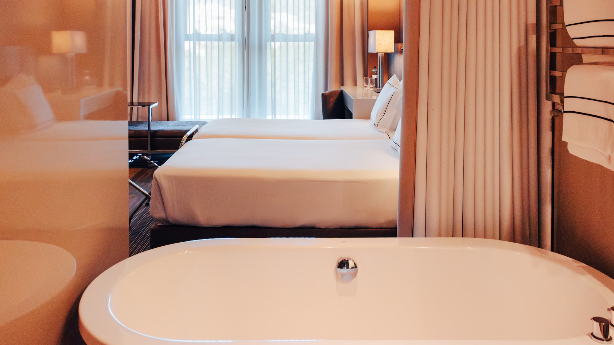 View from white bathtub into a luxury hotel room with twin beds and window.