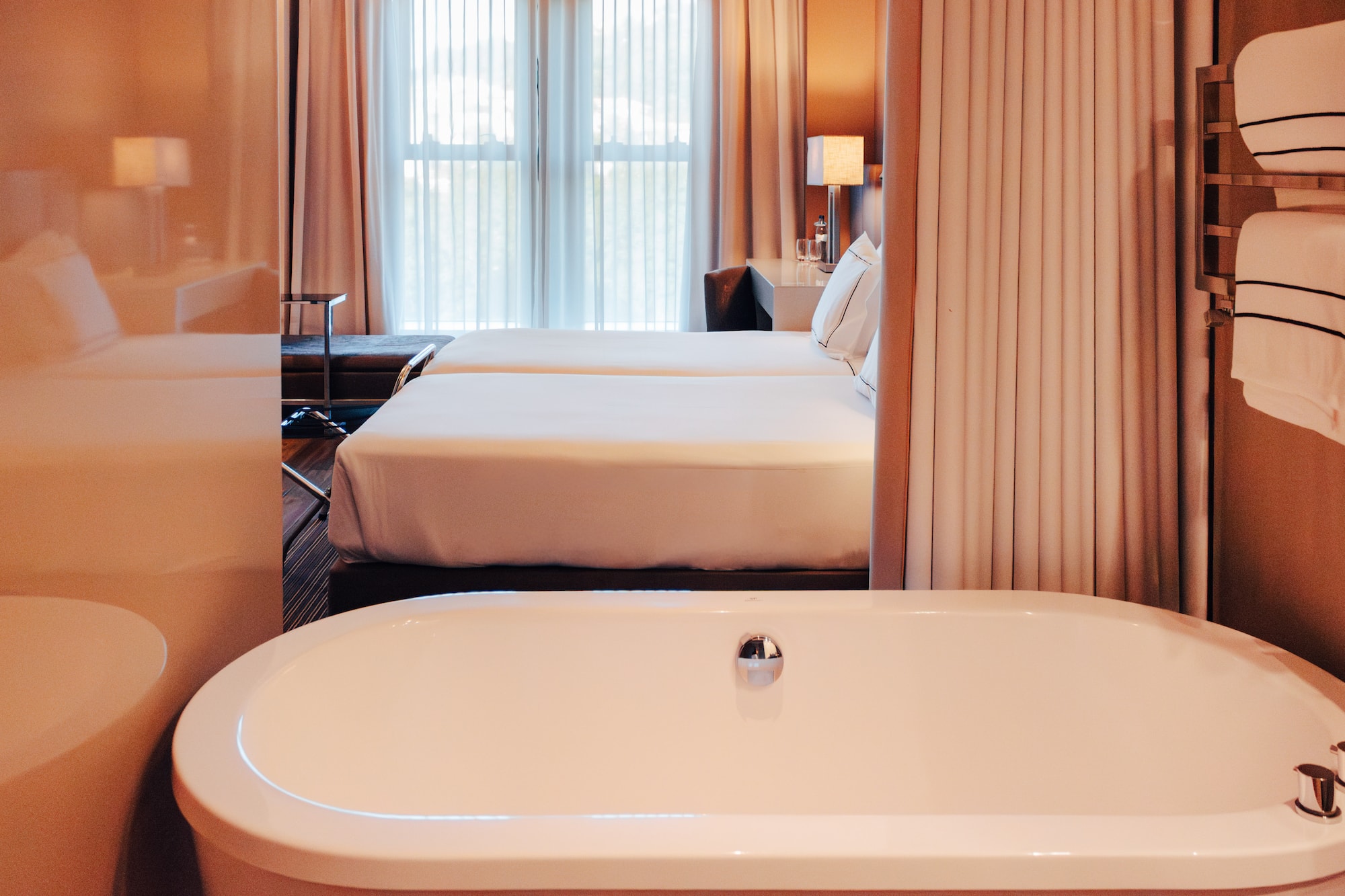 View from white bathtub into a luxury hotel room with twin beds and window.
