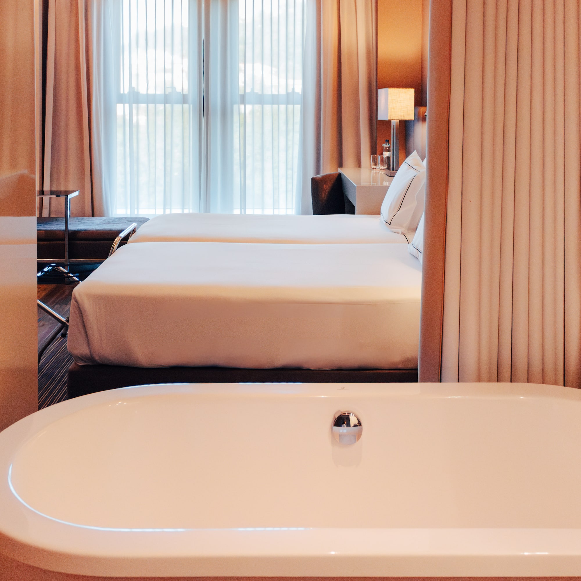View from white bathtub into a luxury hotel room with twin beds and window.