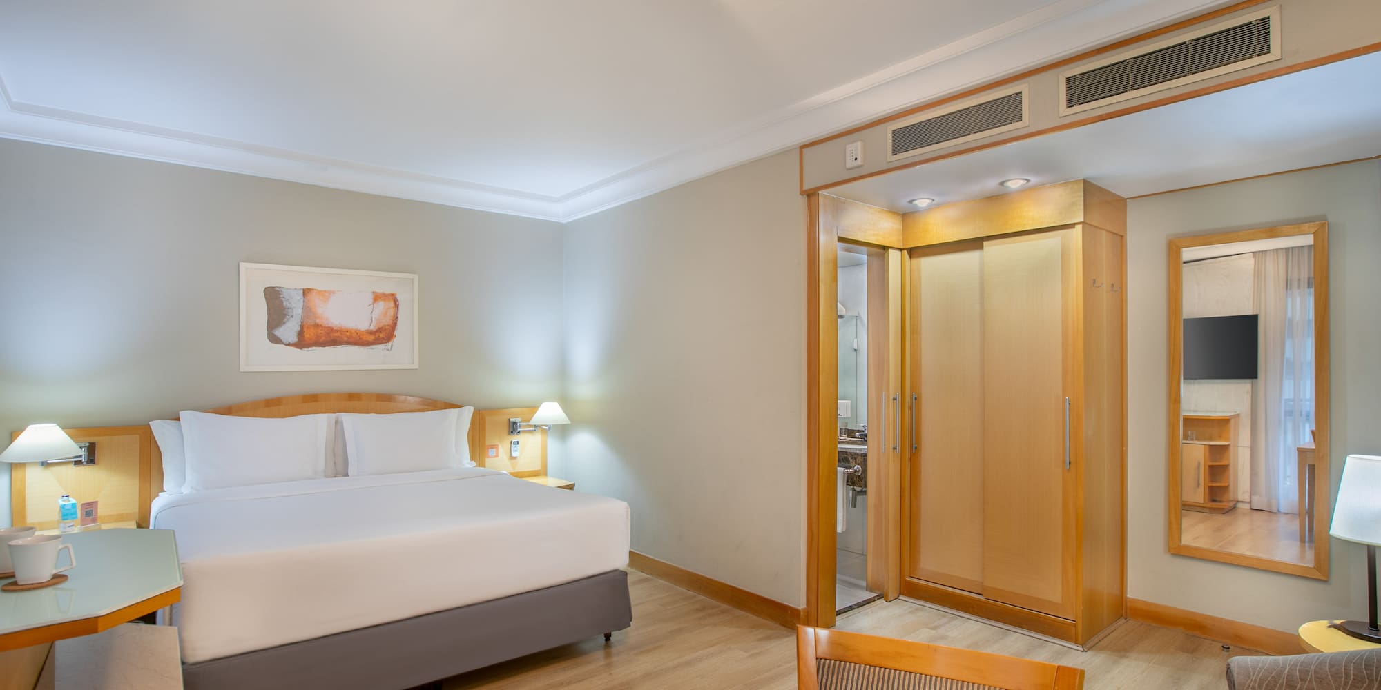 Modern hotel room with king bed, wood floor, desk, wardrobe, and bathroom access.
