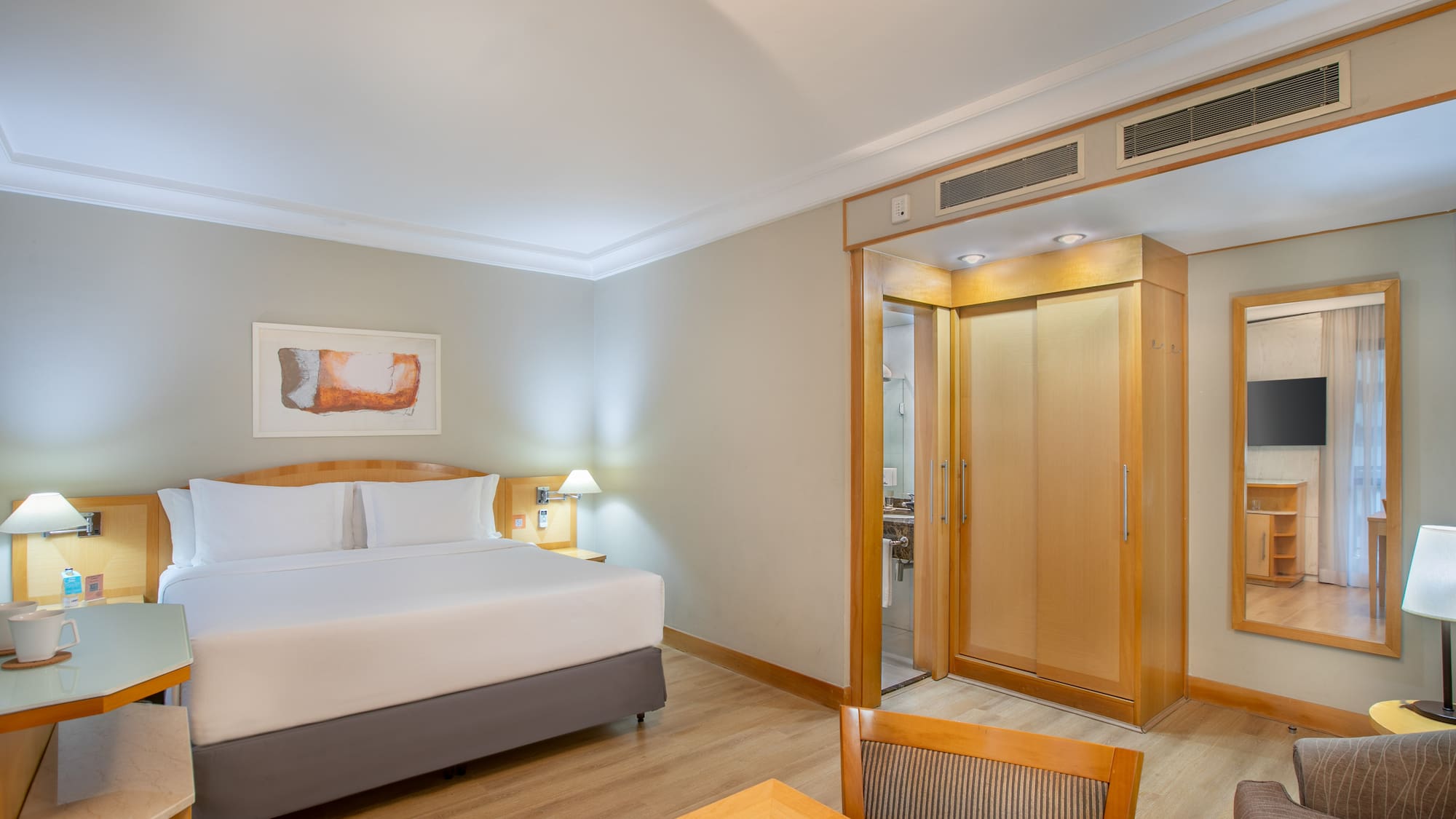 Modern hotel room with king bed, wood floor, desk, wardrobe, and bathroom access.