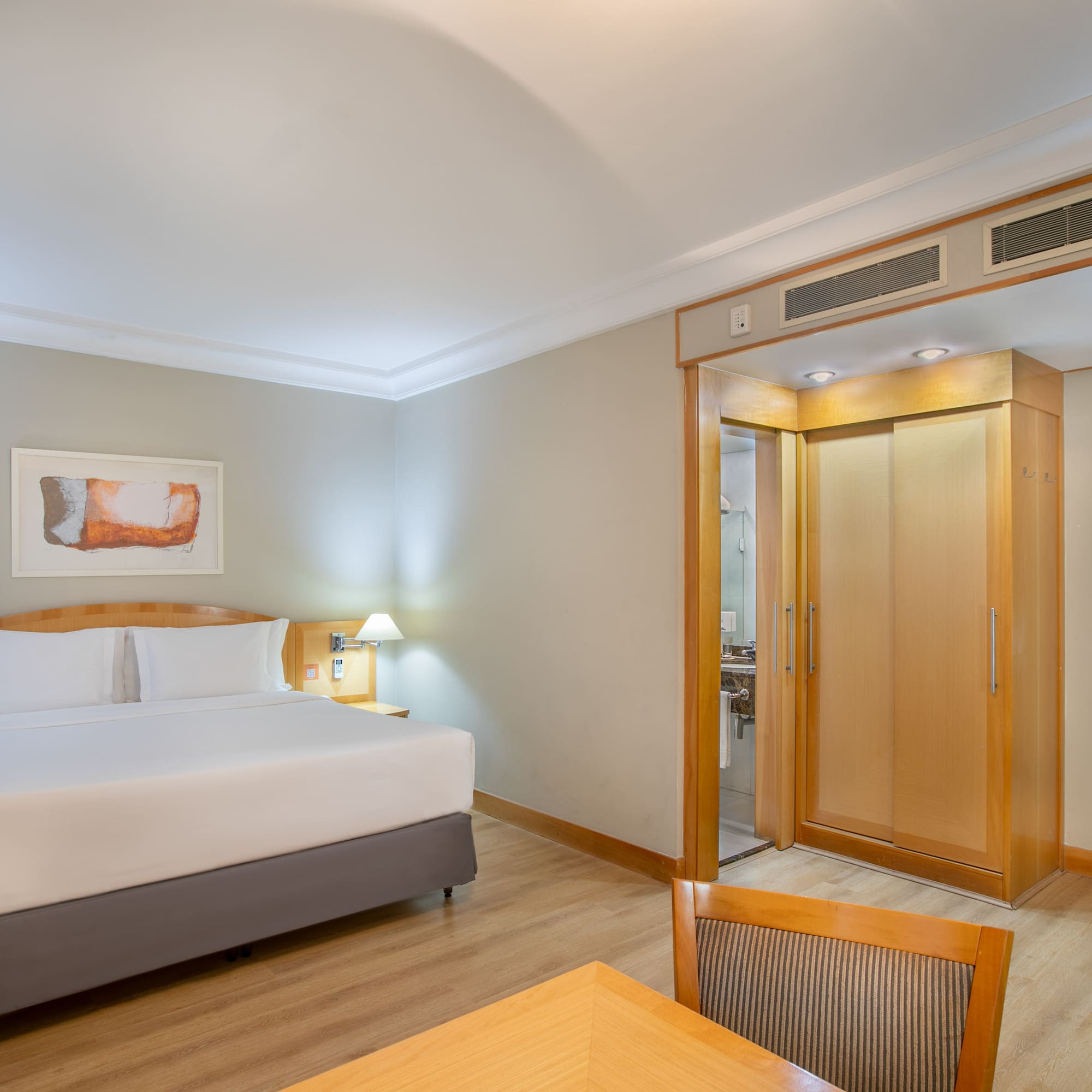 Modern hotel room with king bed, wood floor, desk, wardrobe, and bathroom access.