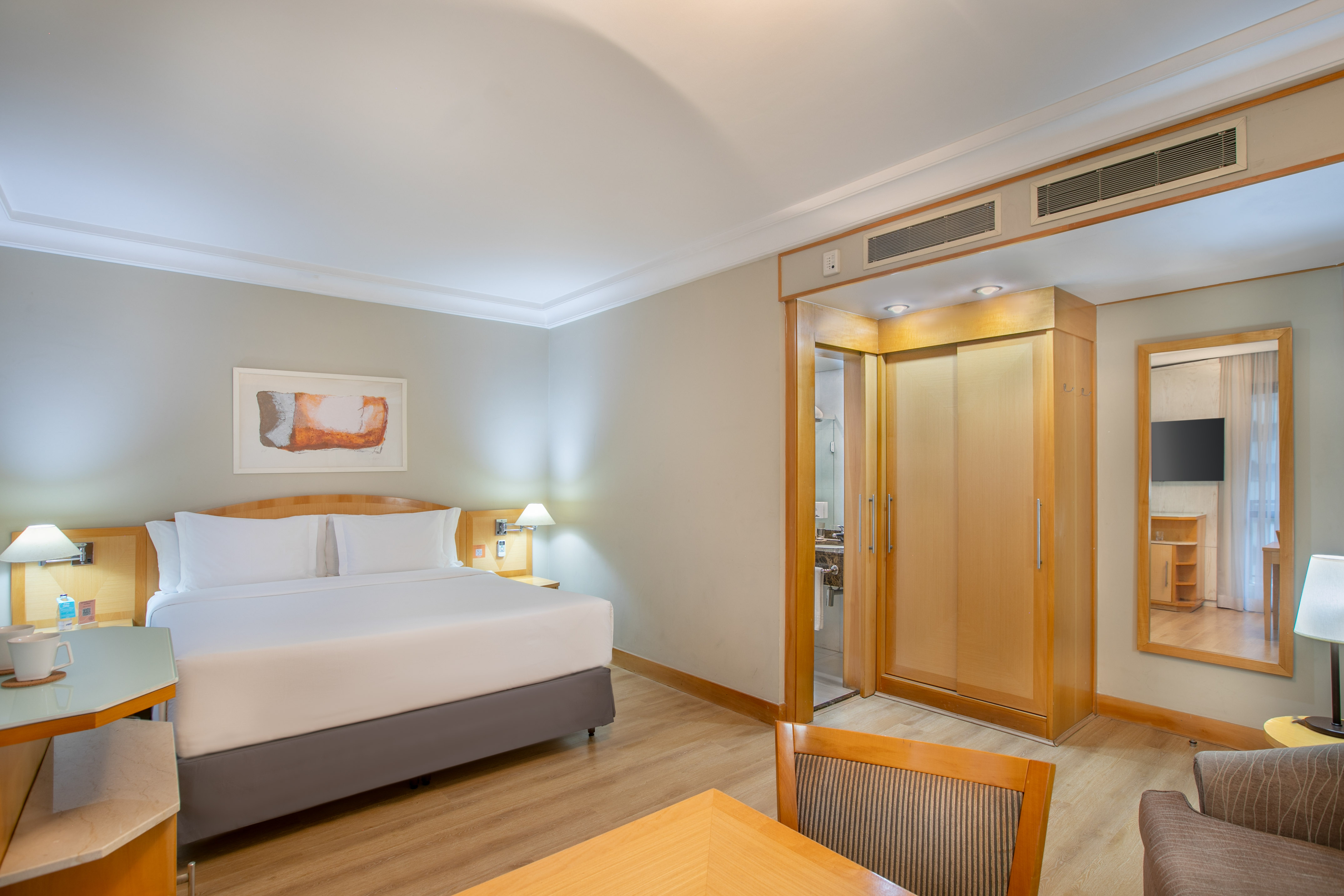 Modern hotel room with king bed, wood floor, desk, wardrobe, and bathroom access.