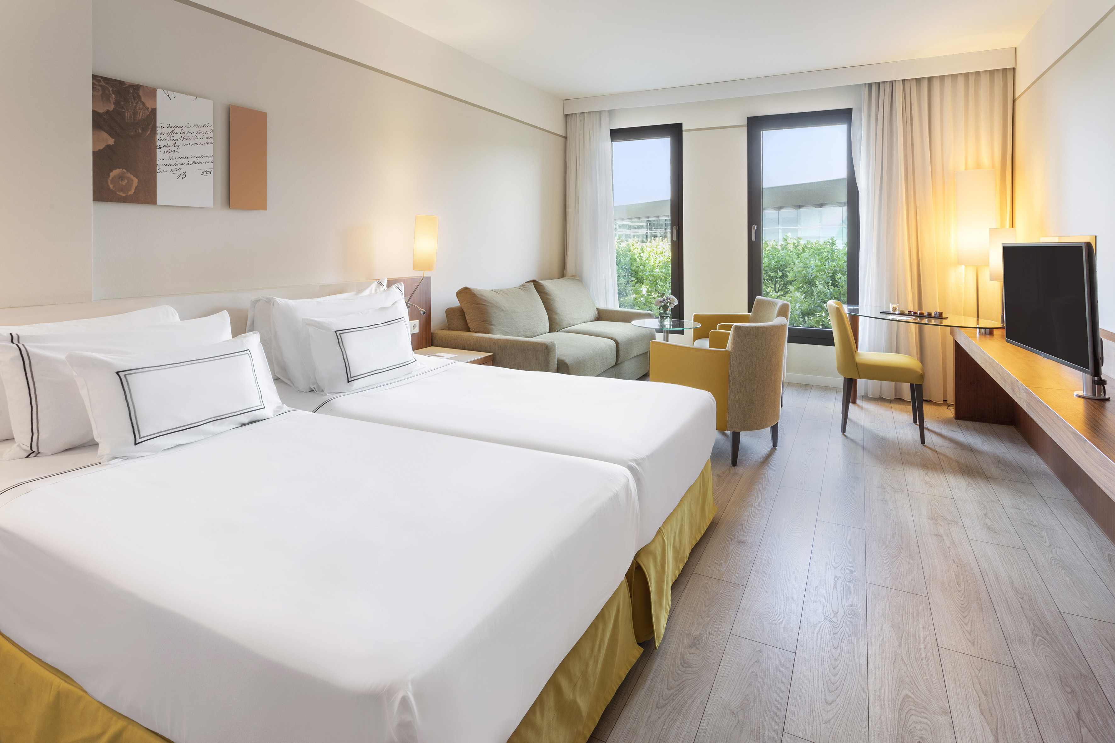 Bright hotel room with twin beds, sofa, desk, TV, wood floor, and windows.
