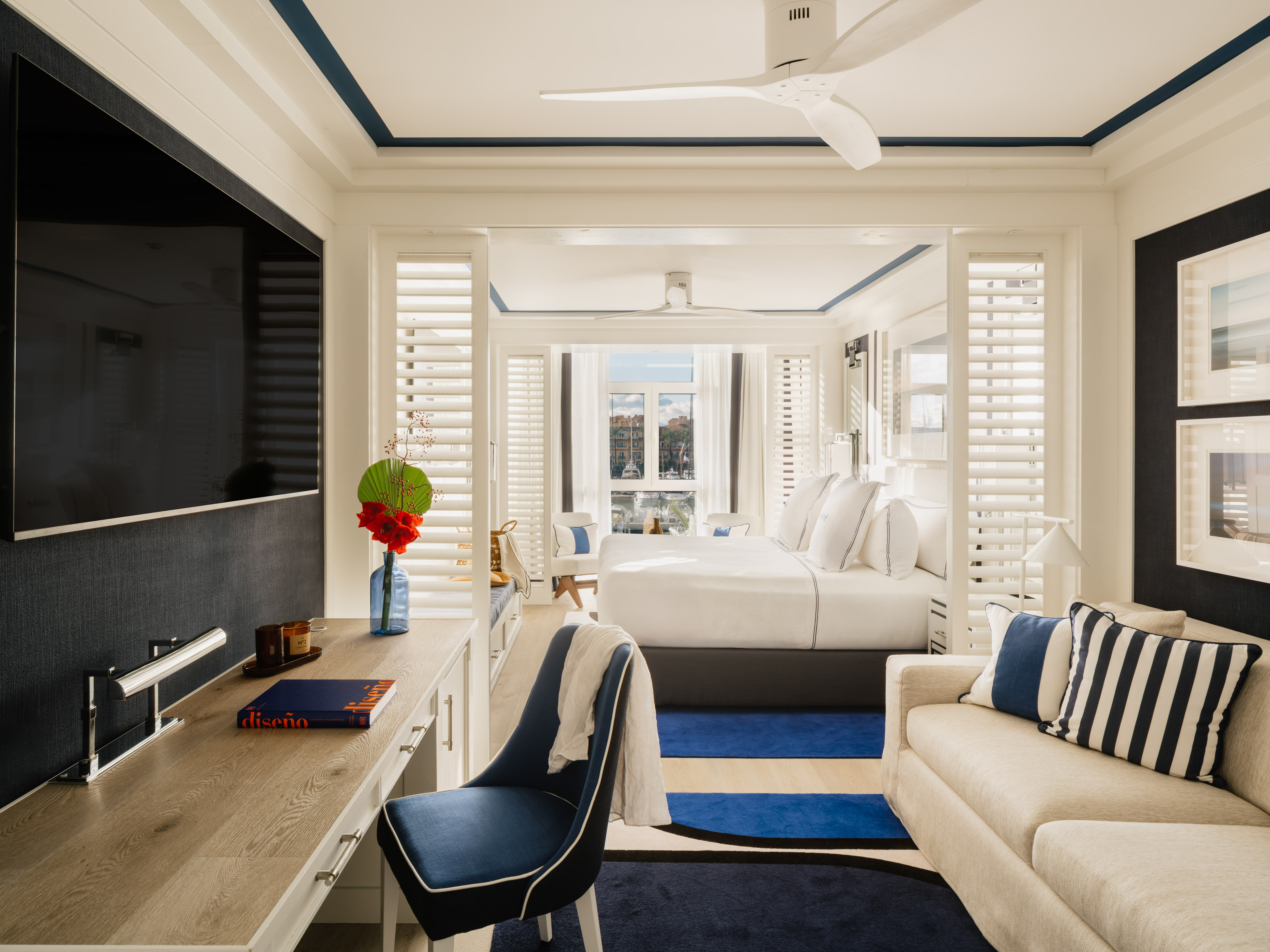Modern blue and white hotel suite with bed, sofa, desk, and water view.