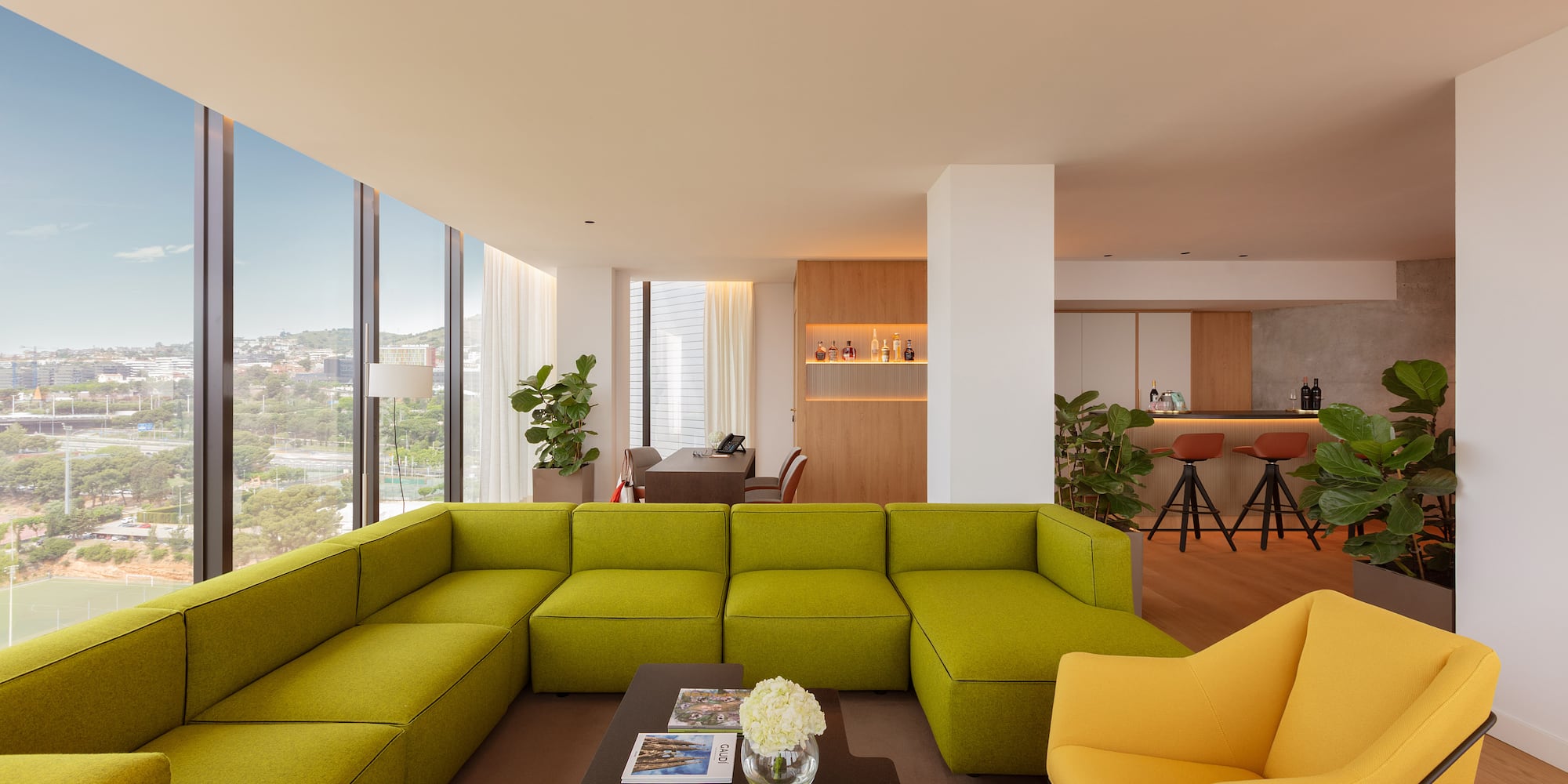 Bright lounge with green sectional, yellow armchairs, bar area, and city view.