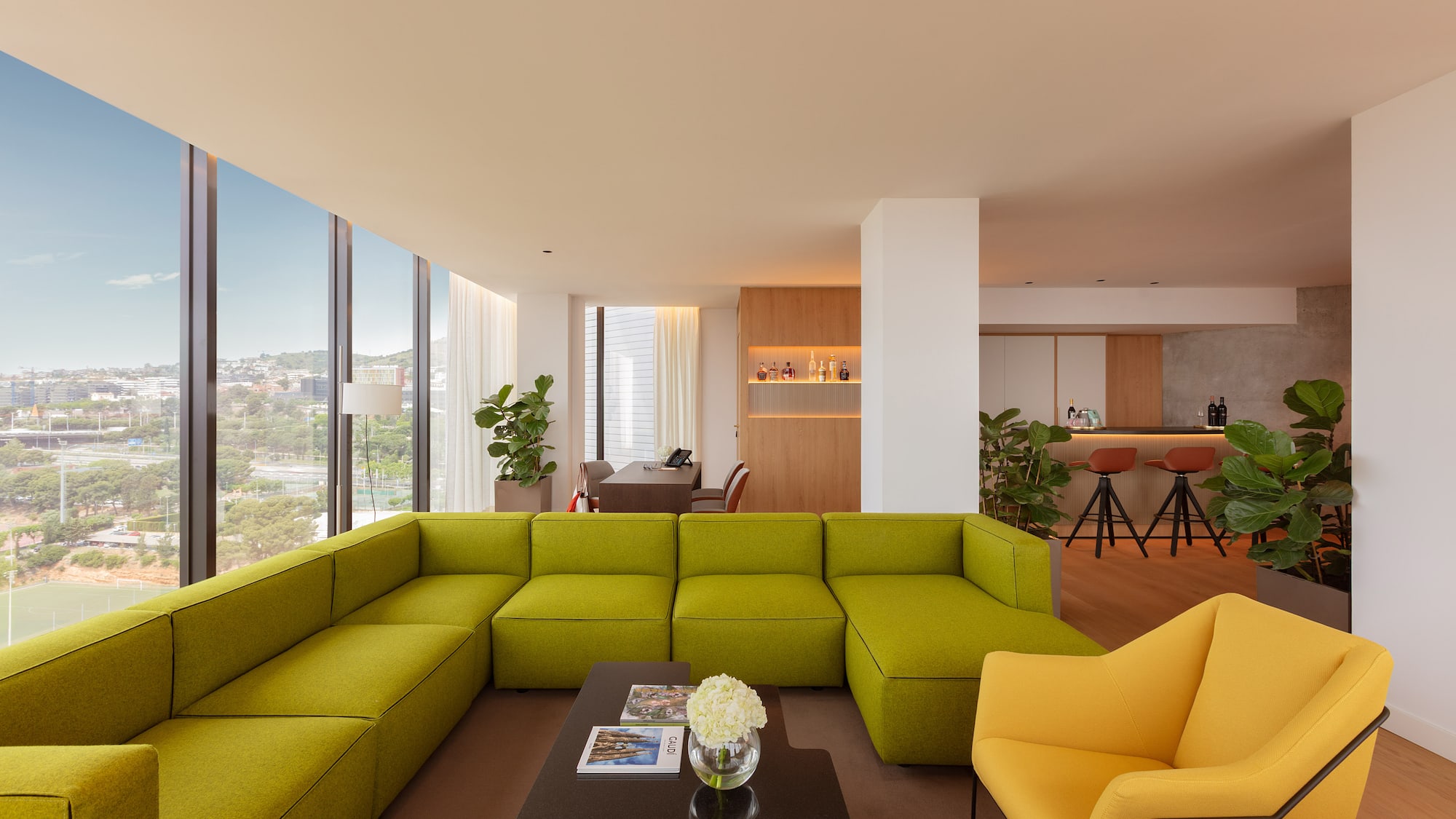 Bright lounge with green sectional, yellow armchairs, bar area, and city view.