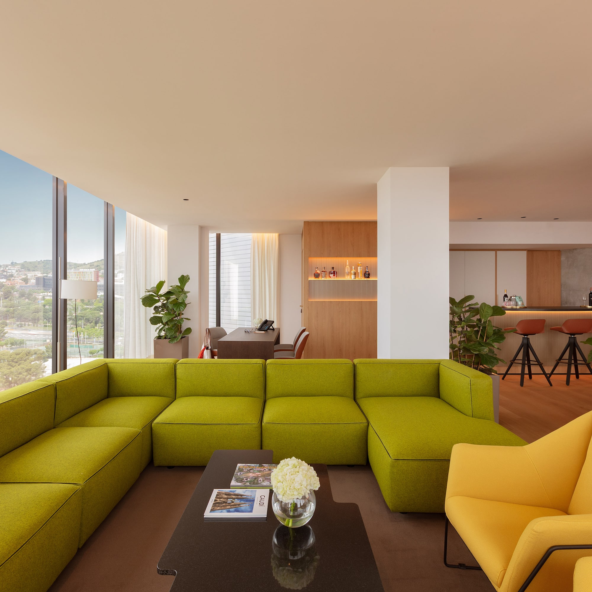 Bright lounge with green sectional, yellow armchairs, bar area, and city view.