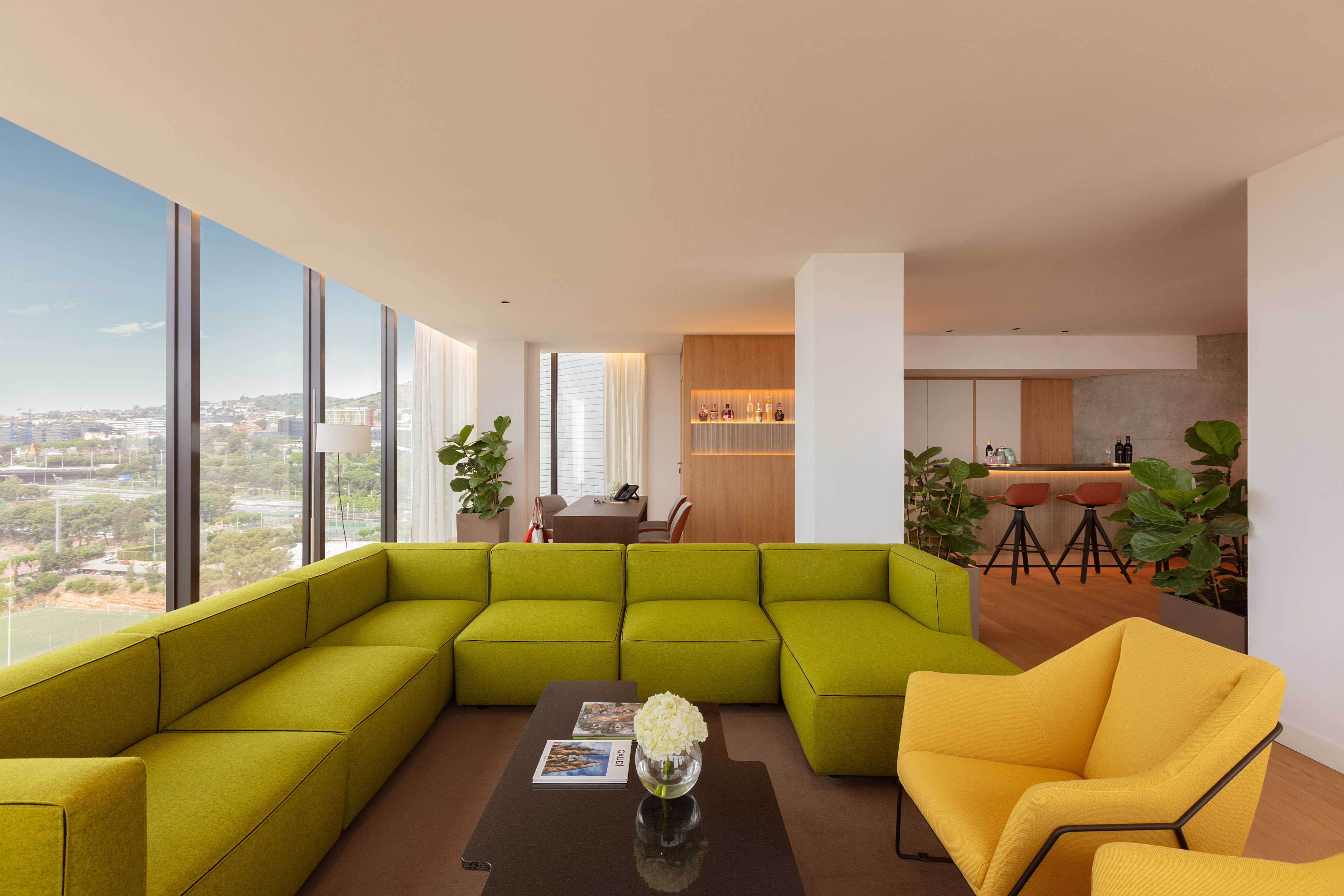 Bright lounge with green sectional, yellow armchairs, bar area, and city view.