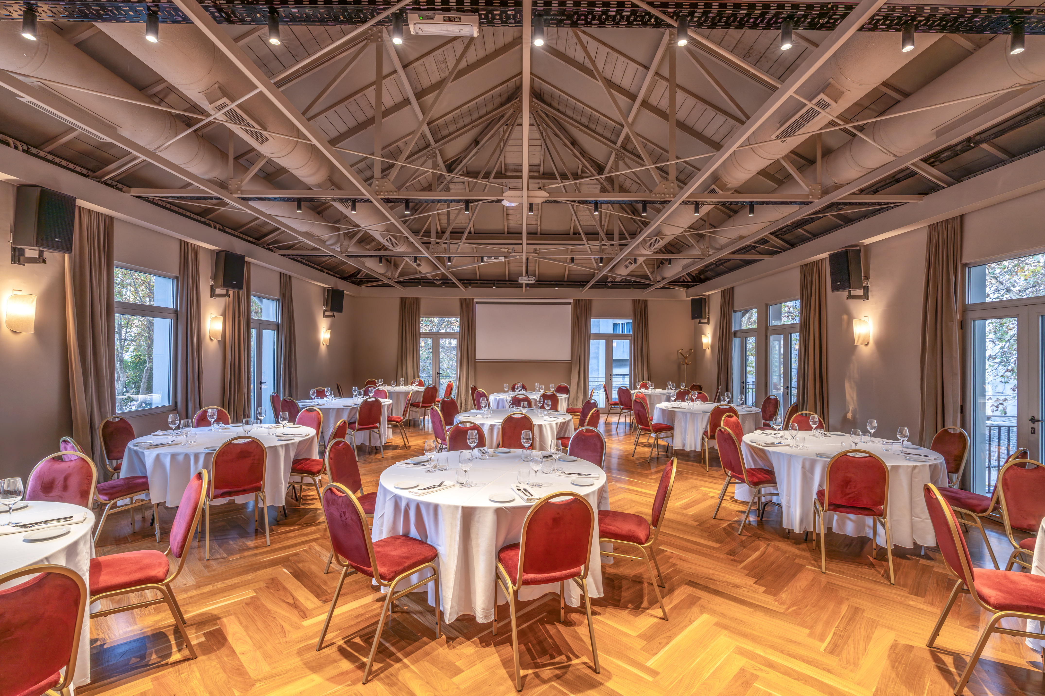 Spacious banquet hall with round tables, red chairs, parquet floor, projector screen.