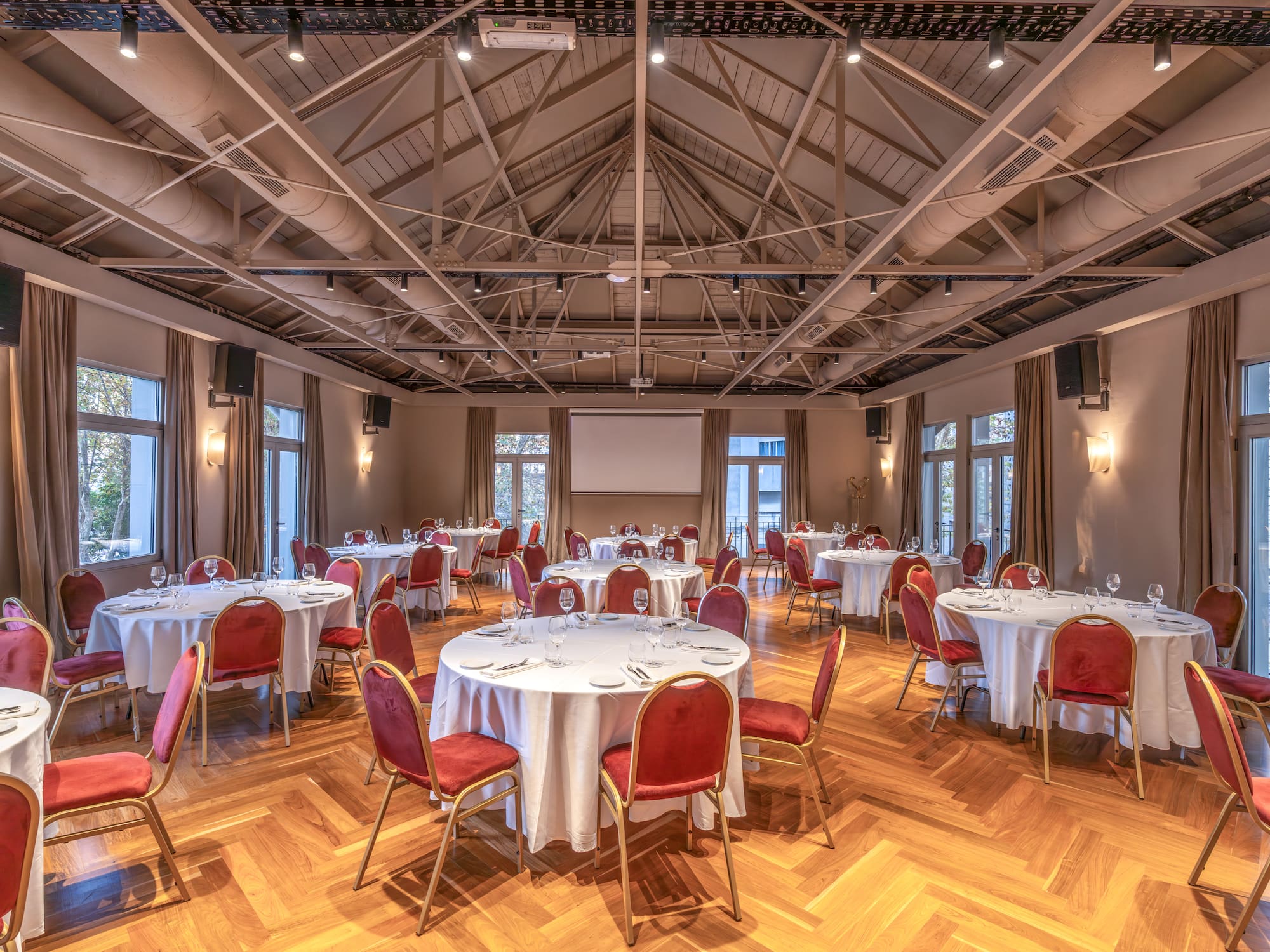 Spacious banquet hall with round tables, red chairs, parquet floor, projector screen.