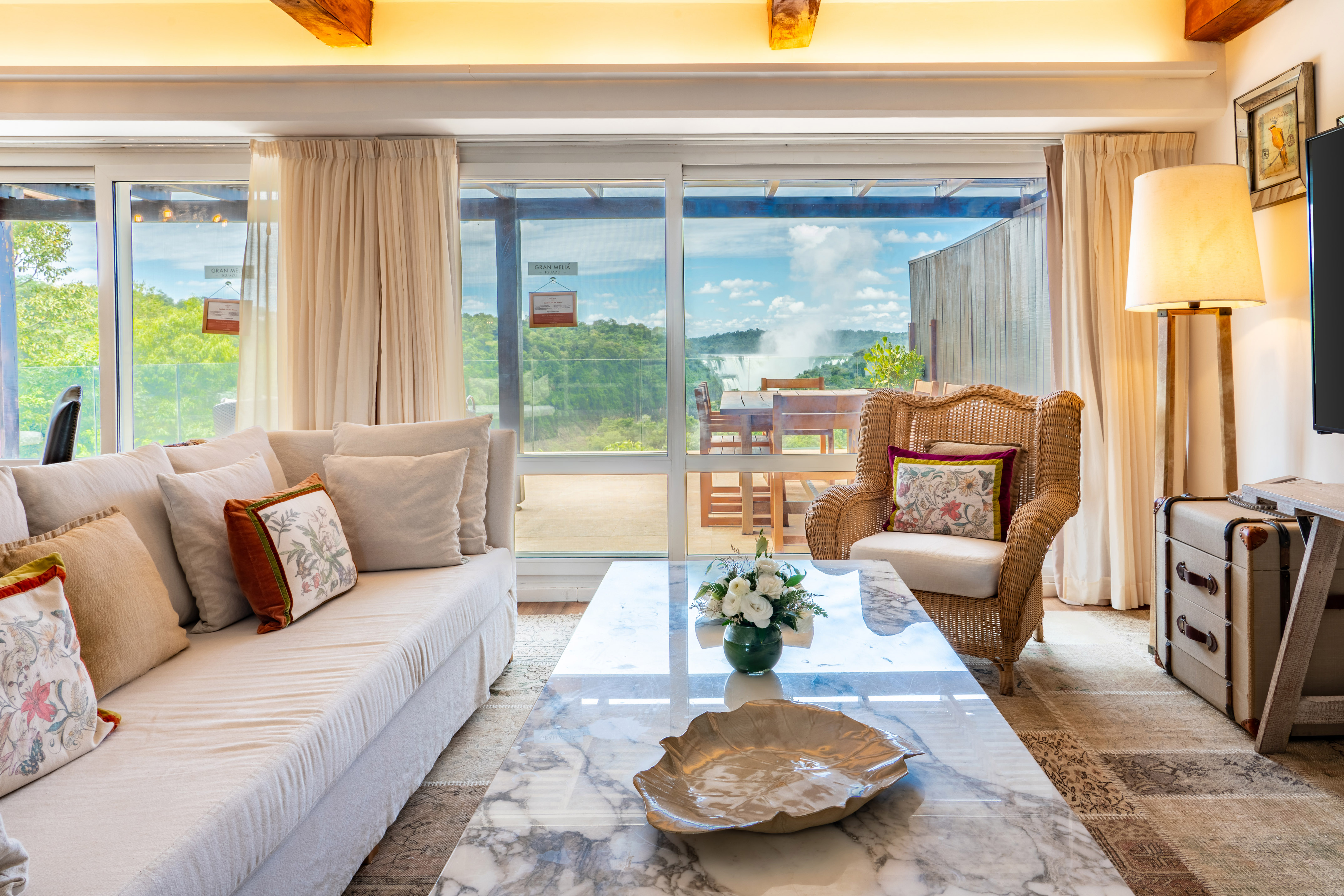 Hotel living room with sofa, wicker chair, marble table, and Iguazu Falls view.