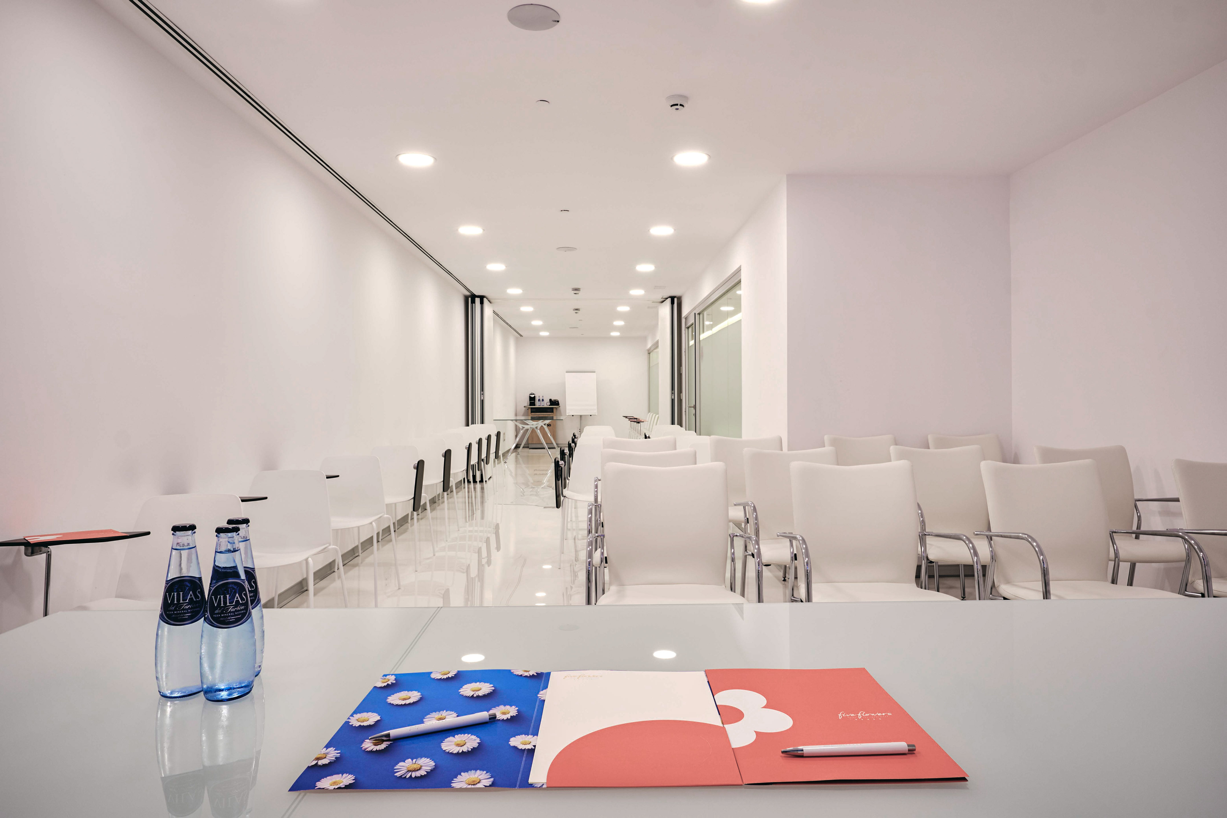 Modern white meeting room with rows of chairs, table, water bottles, and folders.