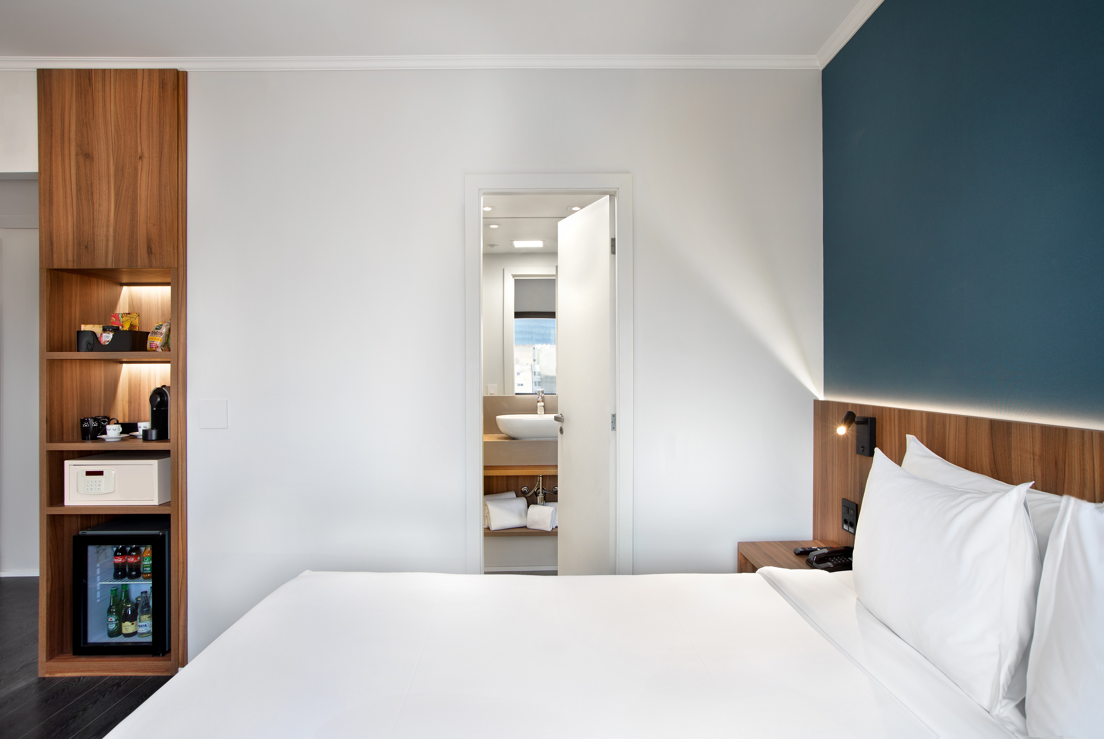 Modern hotel room with white bed, wooden minibar, and open private bathroom.