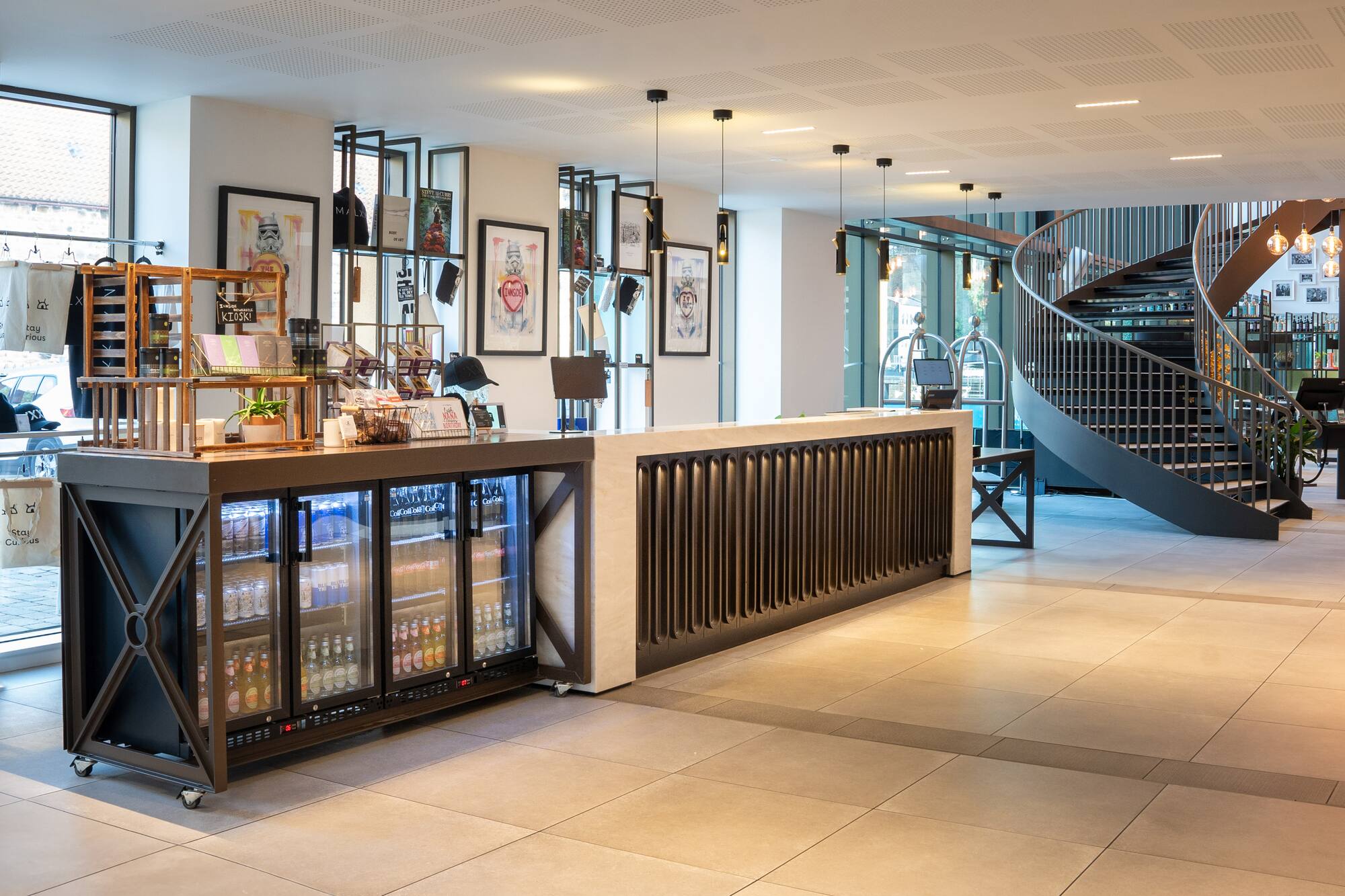 Modern hotel lobby with reception desk, beverage kiosk, and spiral staircase.