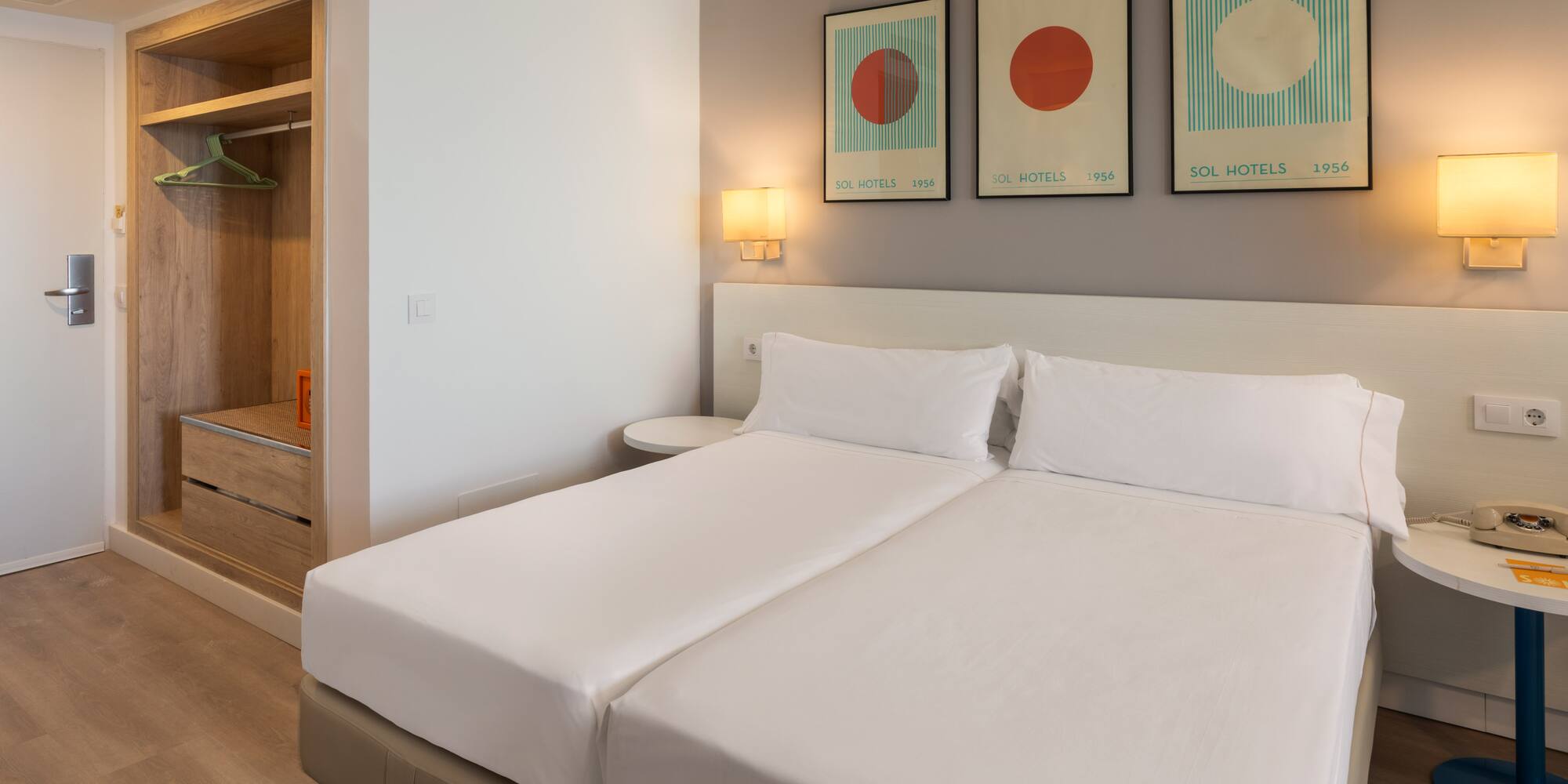 Modern hotel room with twin beds, white bedding, Sol Hotels abstract art, and wooden wardrobe.