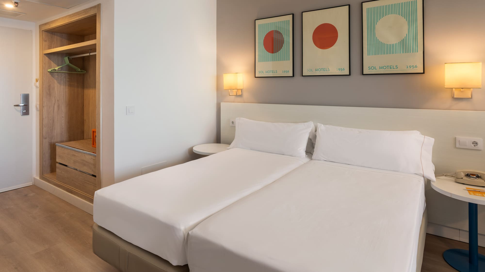 Modern hotel room with twin beds, white bedding, Sol Hotels abstract art, and wooden wardrobe.