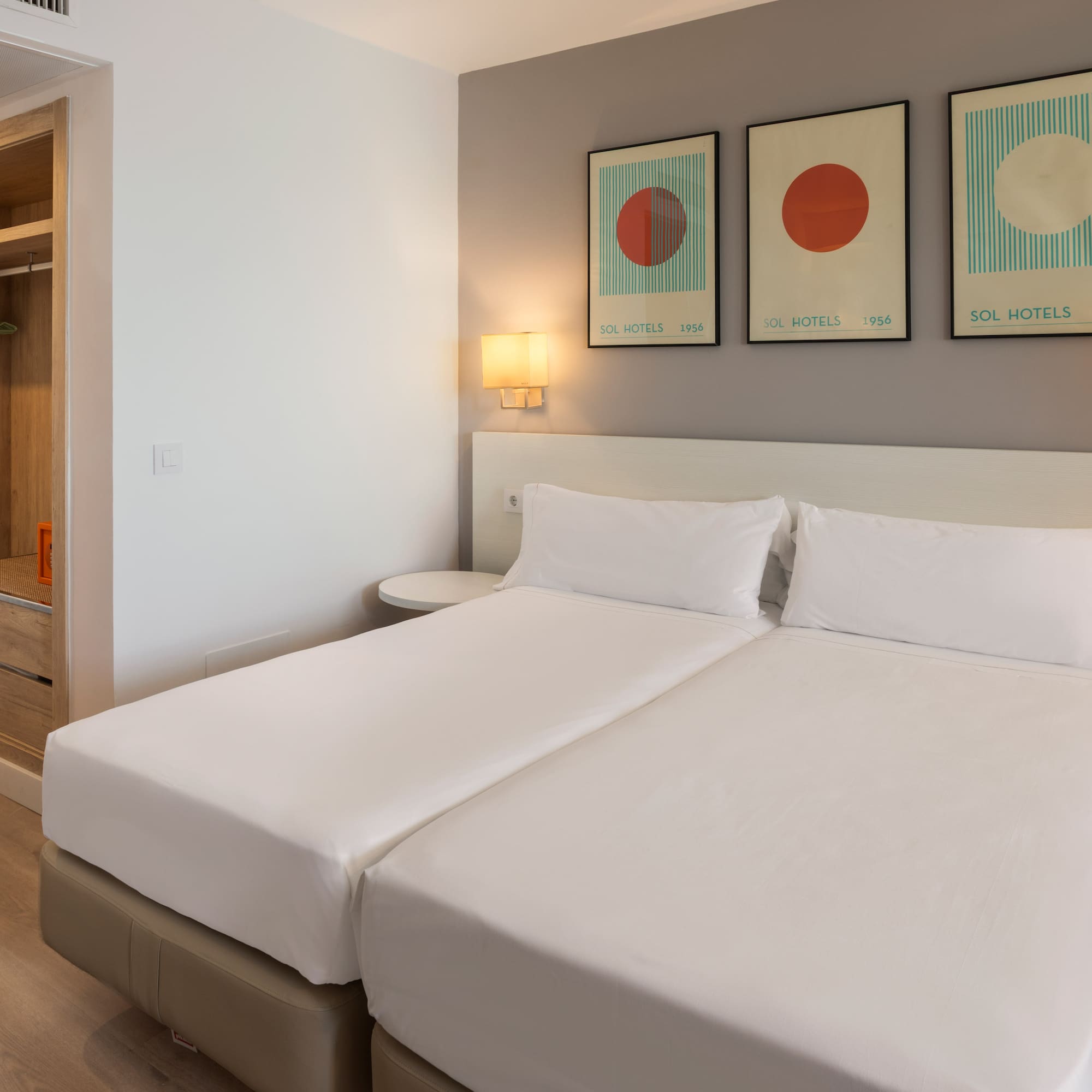 Modern hotel room with twin beds, white bedding, Sol Hotels abstract art, and wooden wardrobe.