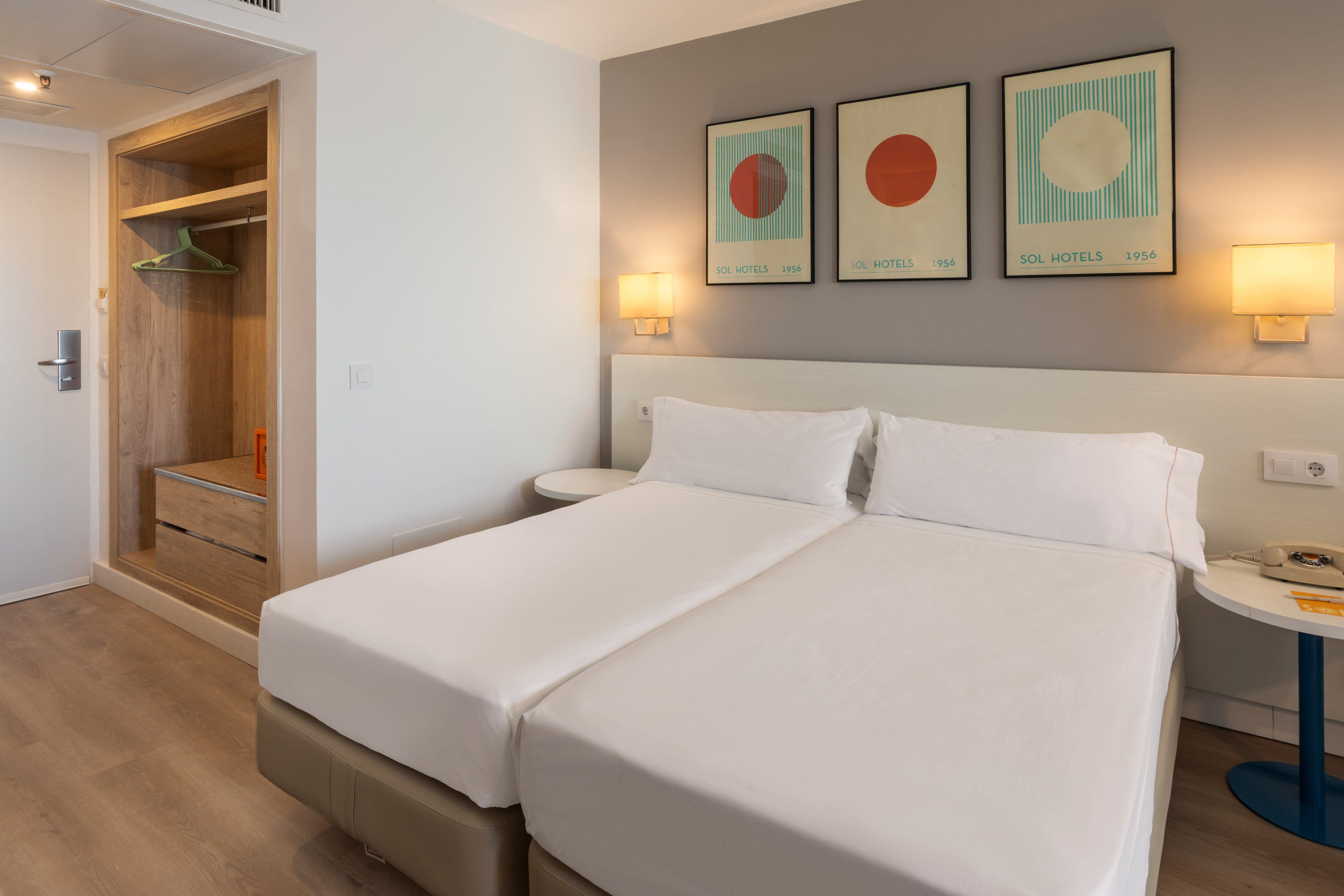 Contemporary hotel room with two single beds, abstract art, and wooden wardrobe.