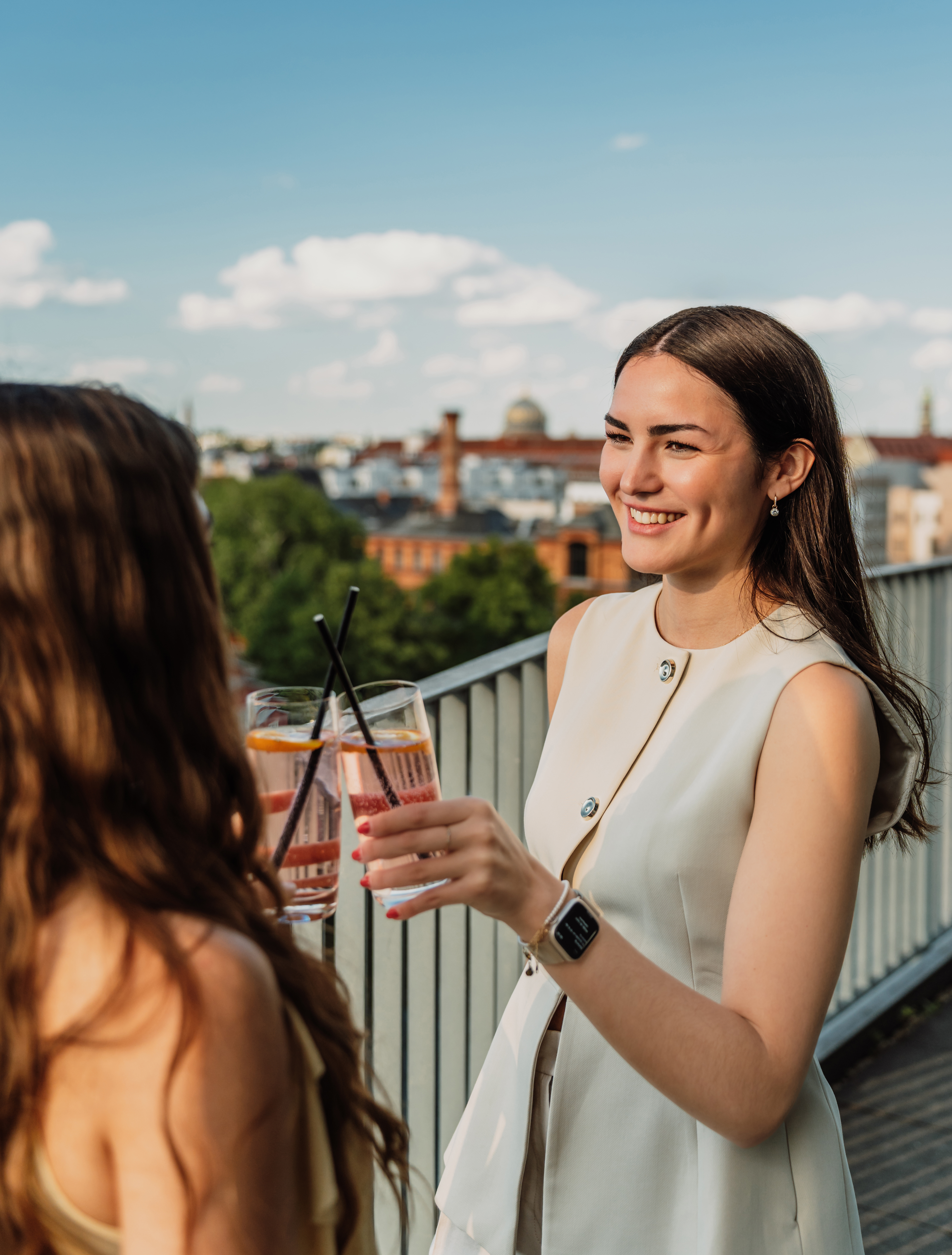 Two smiling women toasting cocktails on a city rooftop with skyline view.