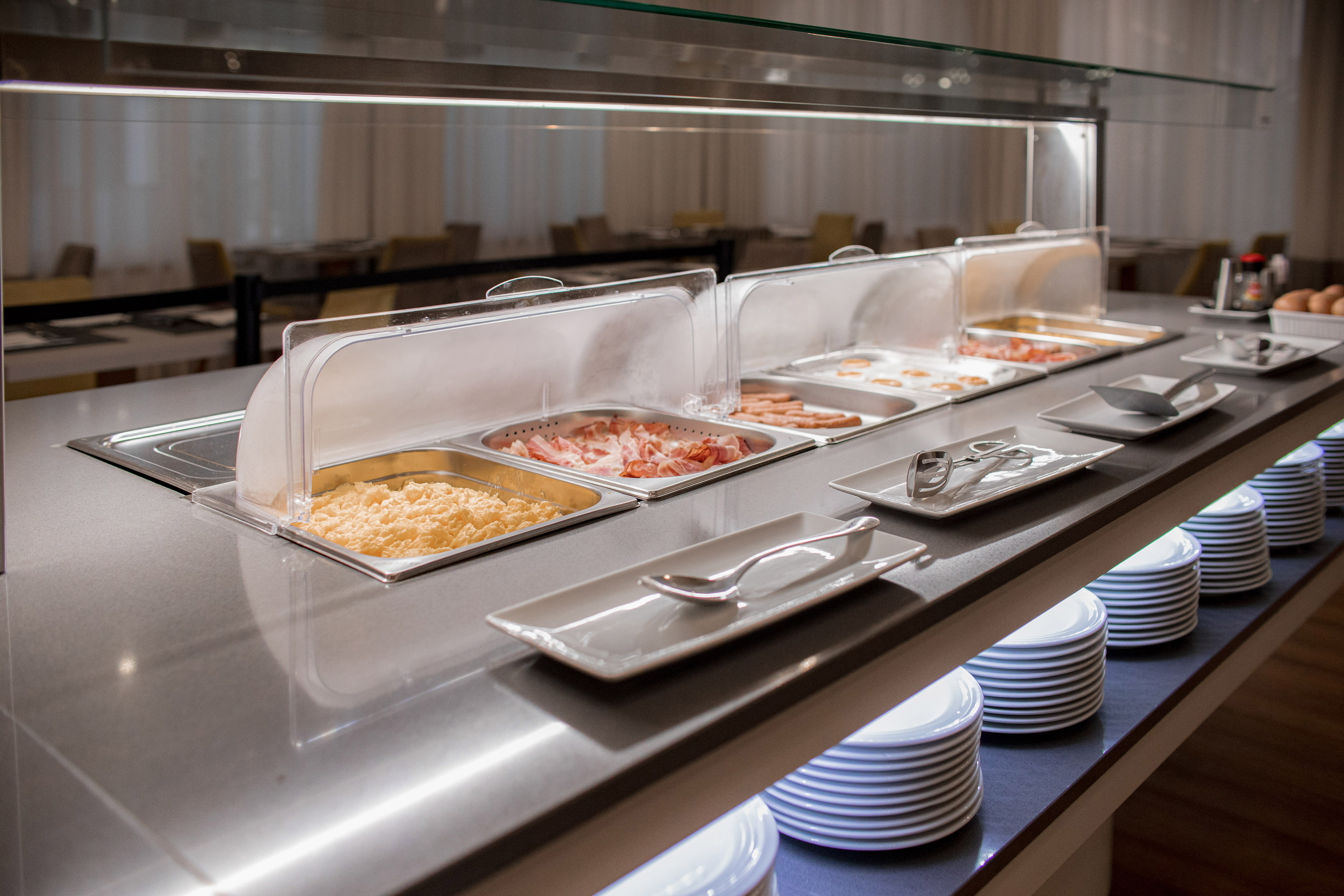 Hotel breakfast buffet with scrambled eggs, bacon, sausages, and plates.