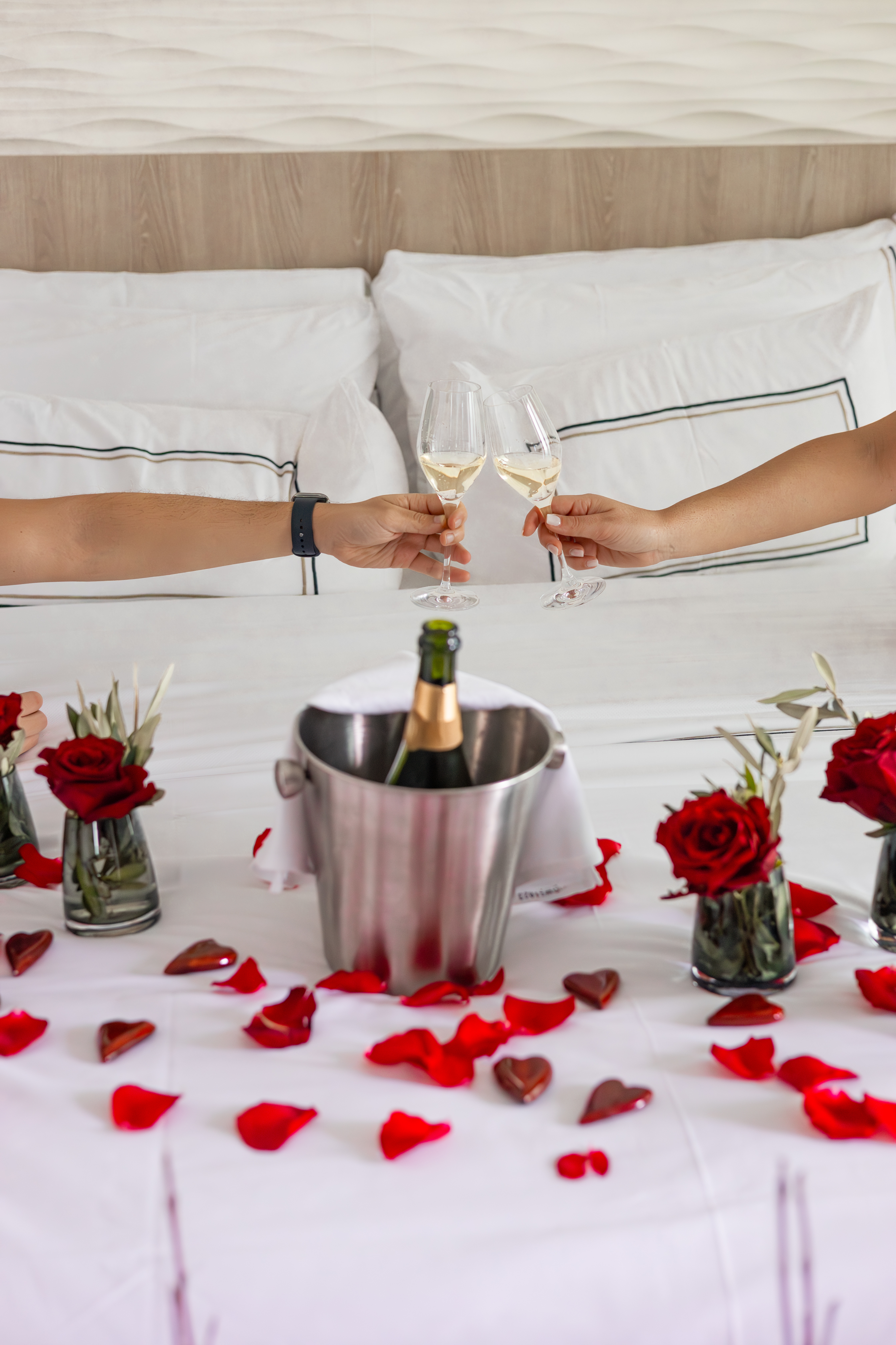 Romantic champagne toast in bed with red rose petals, roses, and chocolates.