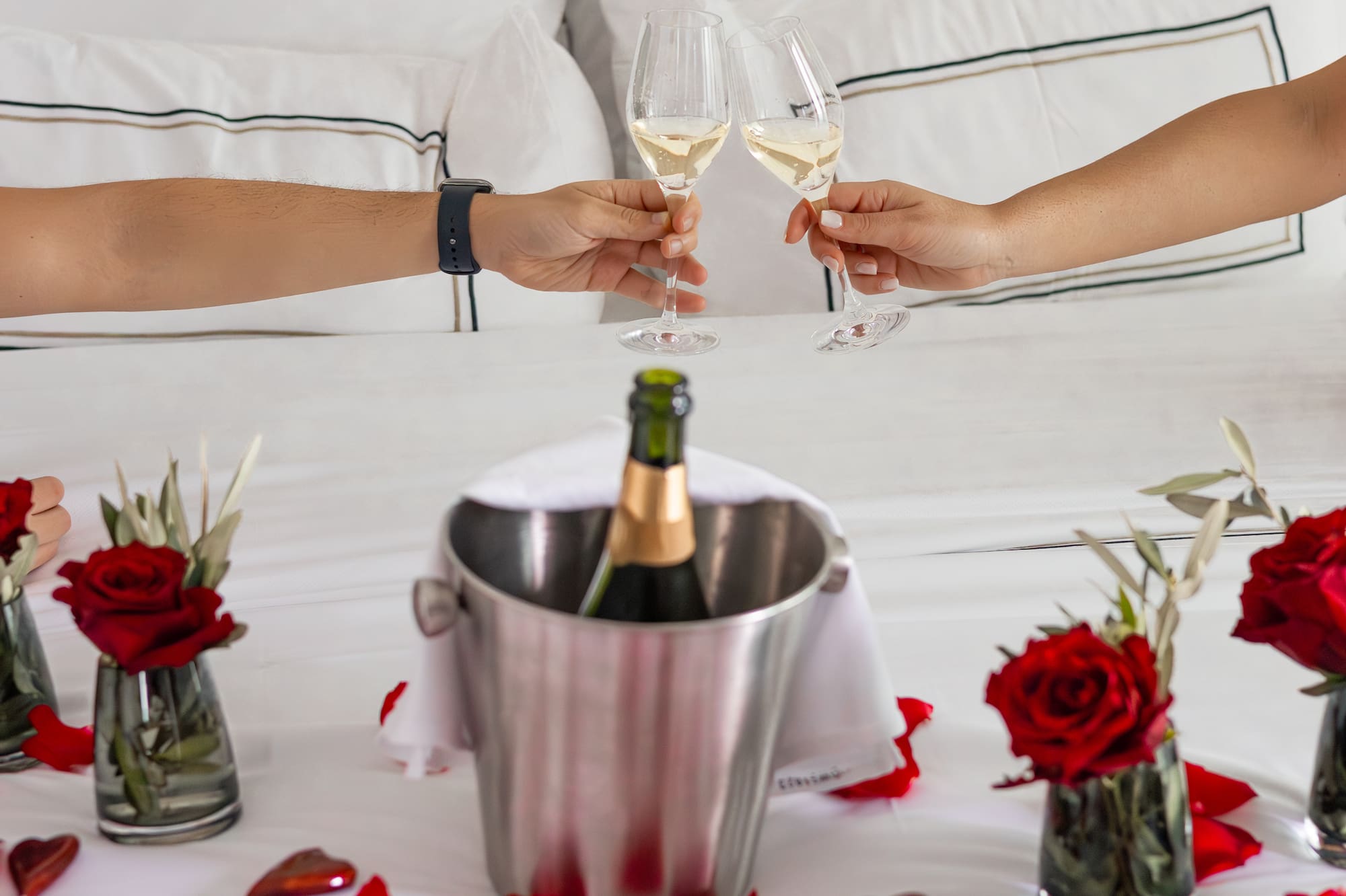 Romantic champagne toast in bed with red rose petals, roses, and chocolates.