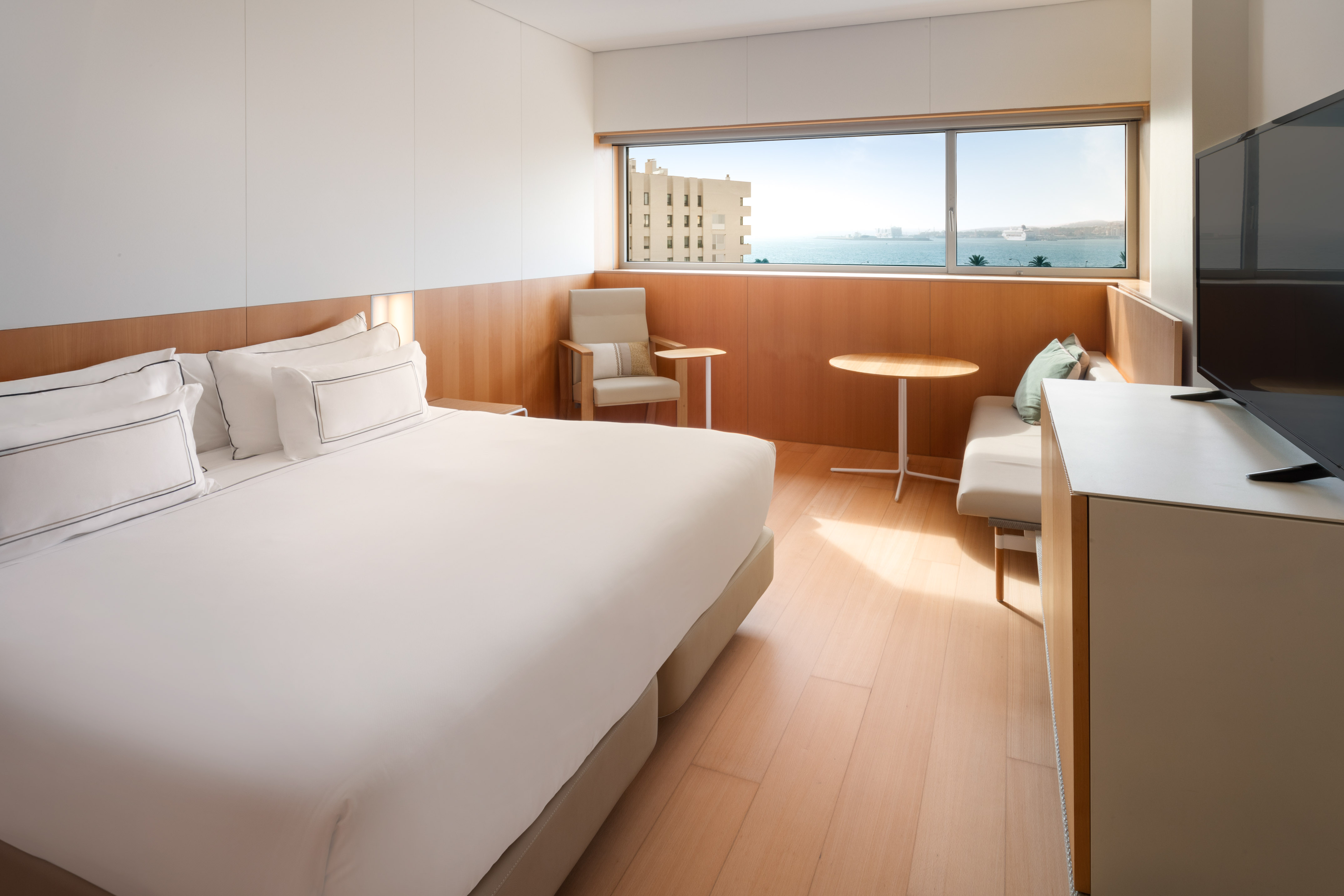 Modern hotel room with sea view, large bed, and light wood decor.