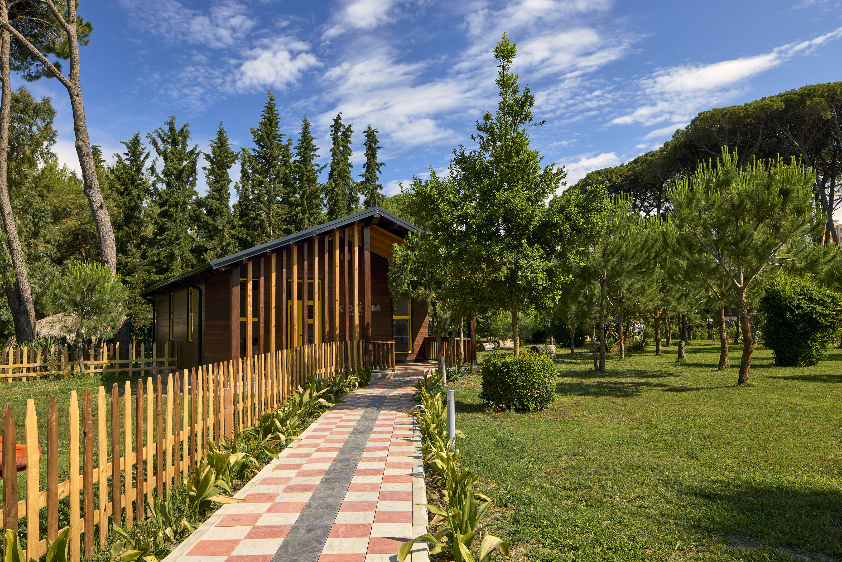 Wooden cabin, checkered path, and picket fence in a green park with pines.