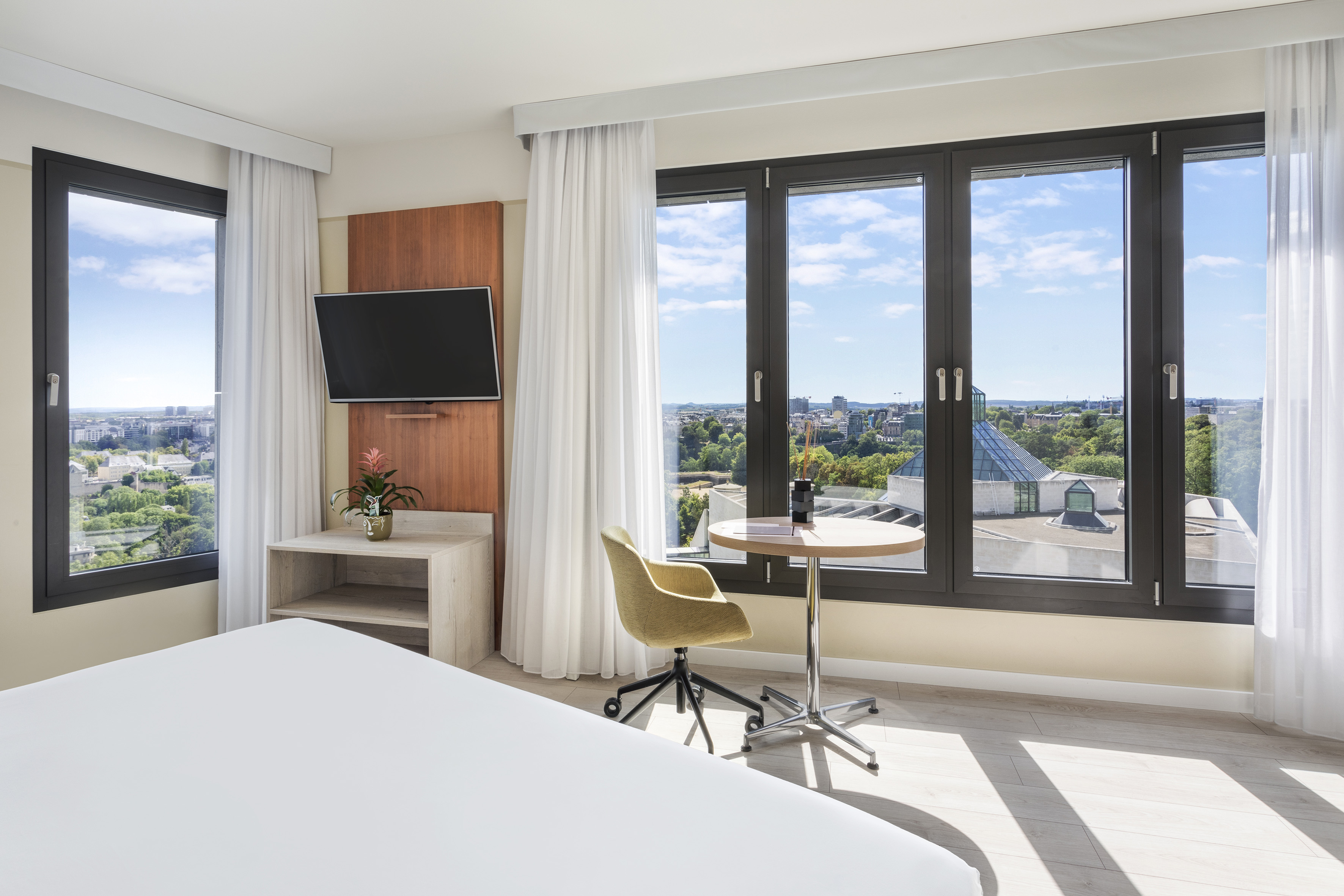 Modern hotel room with city view, bed, TV, and work desk.