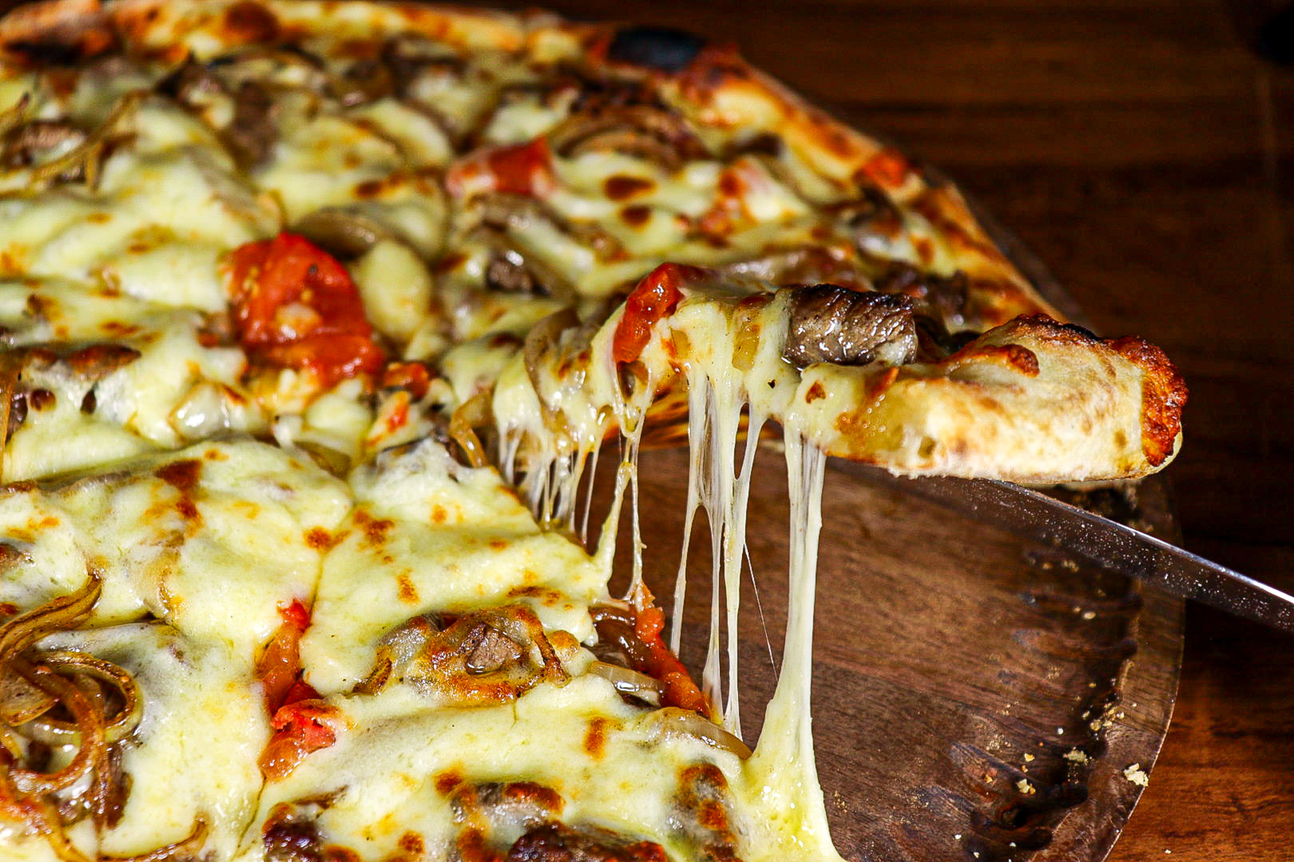 Pizza slice with gooey melted cheese, meat, onions, tomato on a wood board.