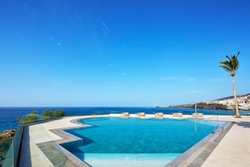 Infinity pool overlooking ocean, with palm tree, coastal village, and blue sky.