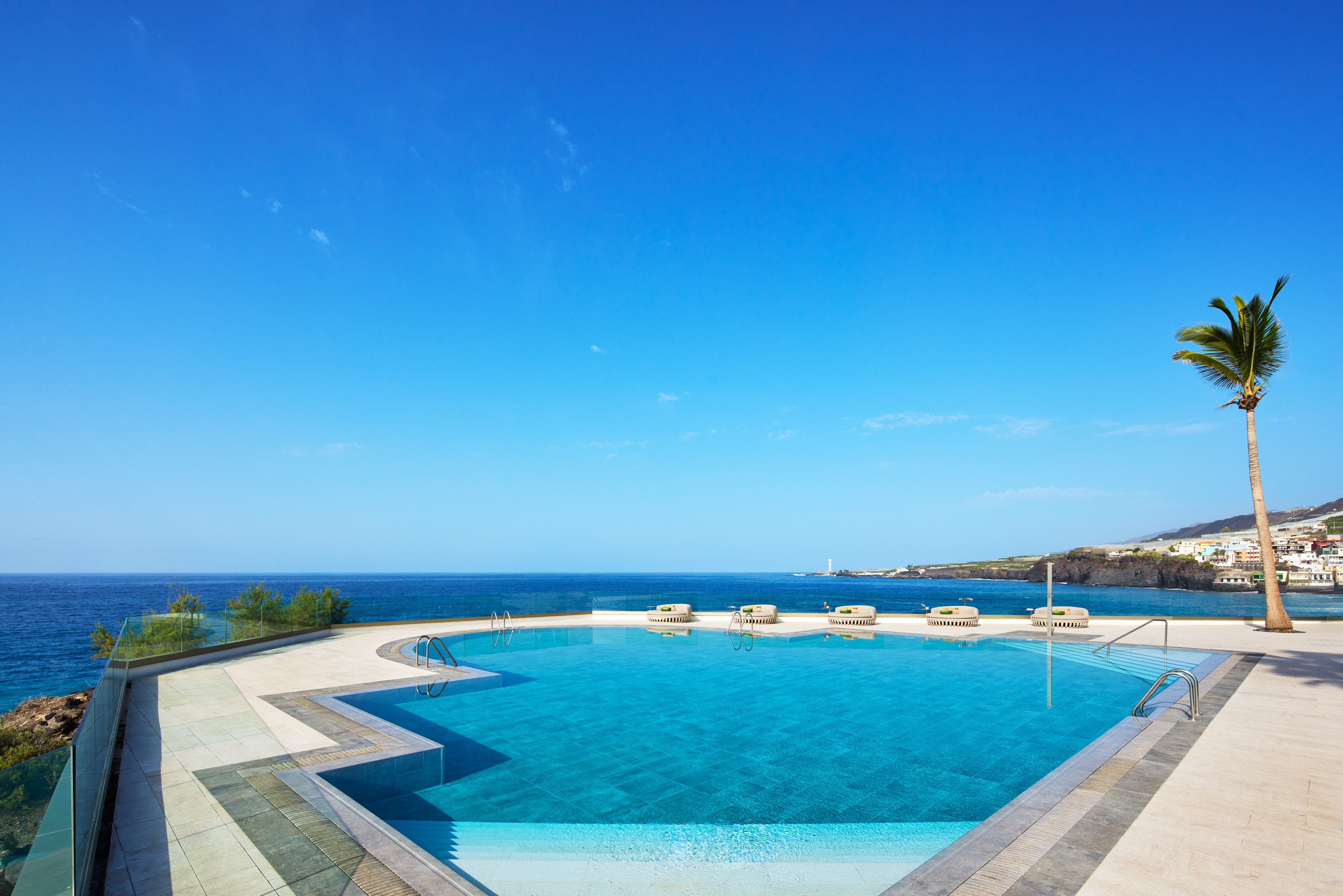 Infinity pool overlooking ocean, with palm tree, coastal village, and blue sky.