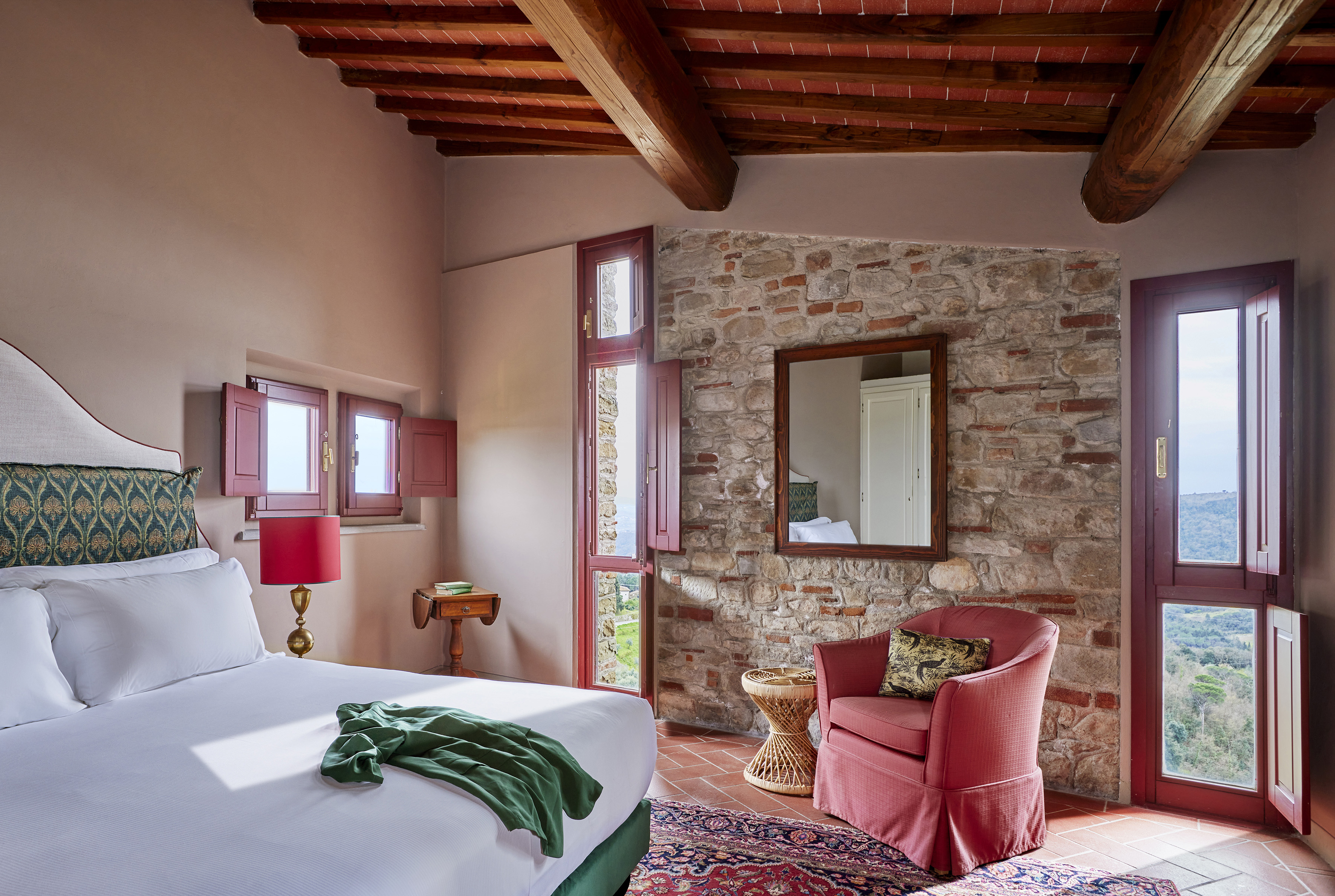 Tuscan-style bedroom with stone wall, beamed ceiling, bed, red armchair, and countryside view.