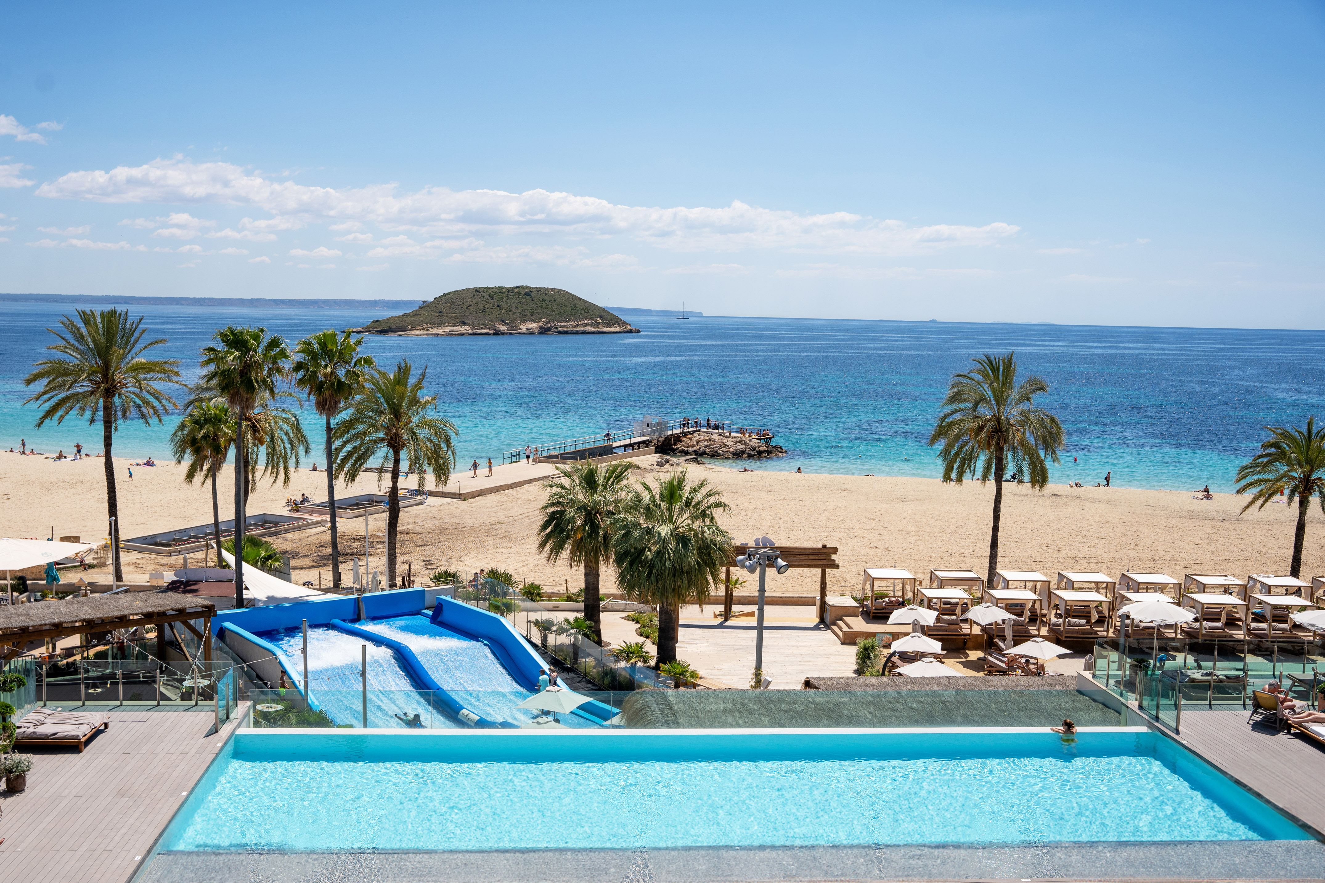 Oceanfront resort with infinity pool, FlowRider, sandy beach, palm trees, and island view.