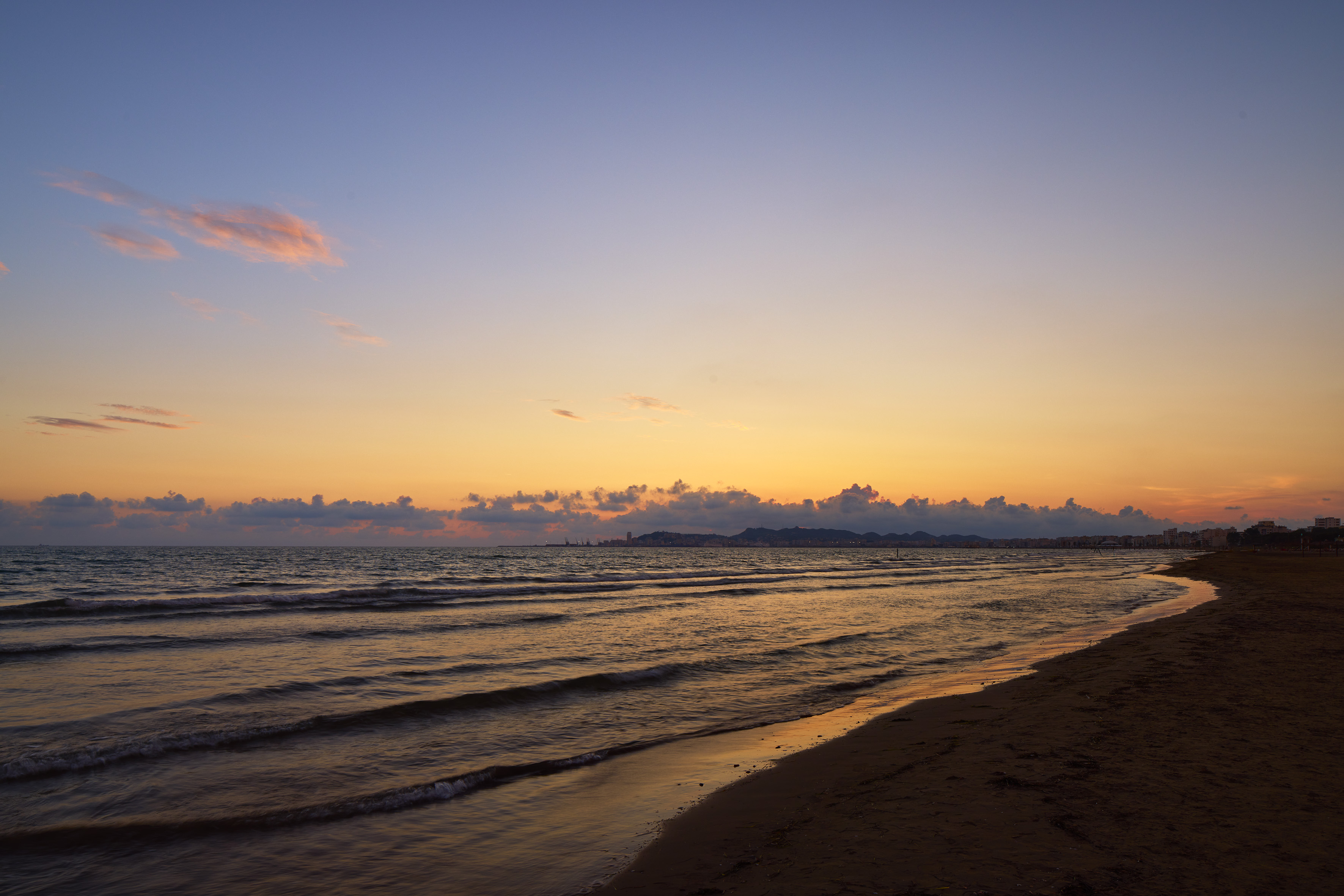 Orange and blue sunset over ocean waves, dark beach, and distant city coastline.