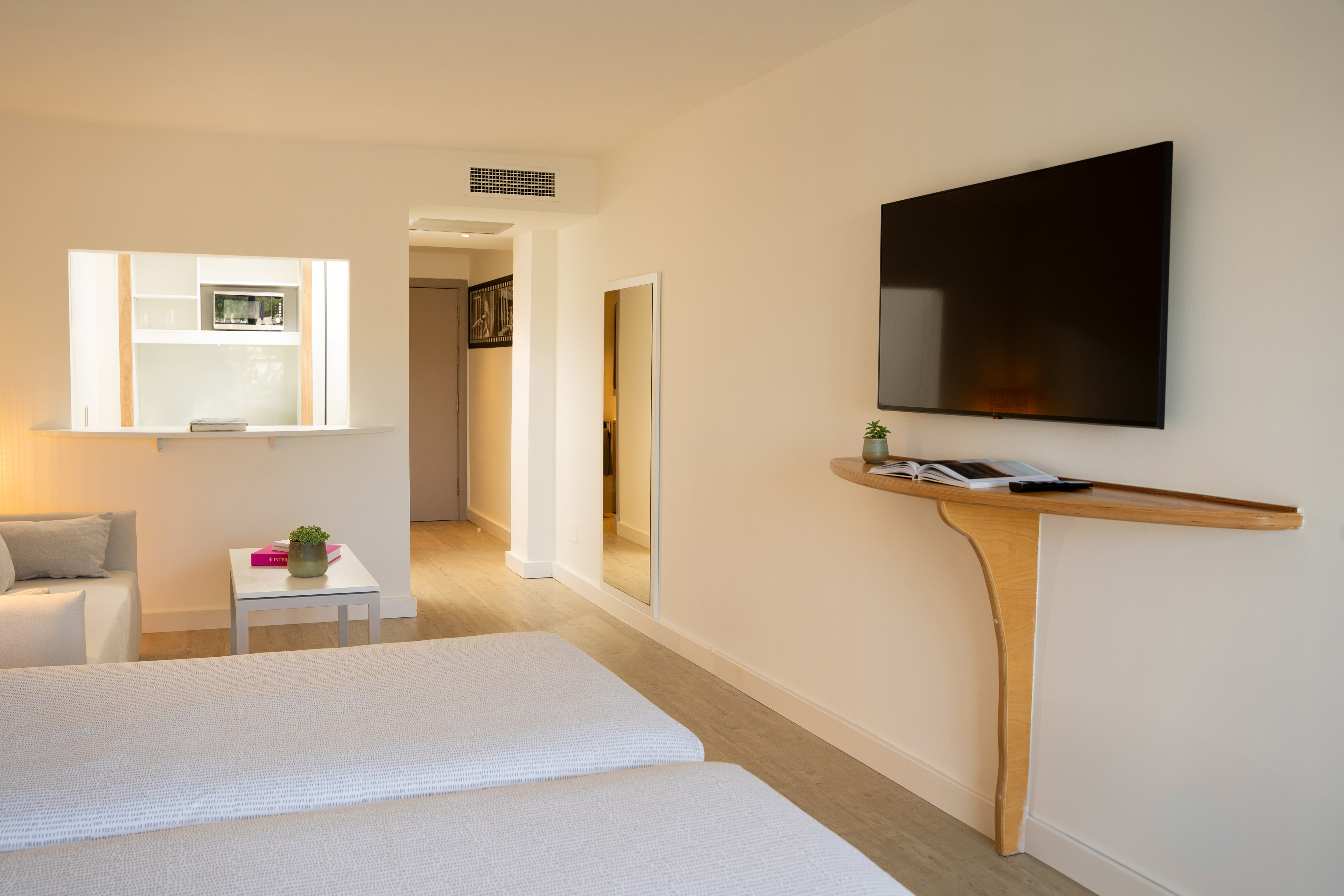 Modern hotel room with two beds, wall TV, and open kitchenette.