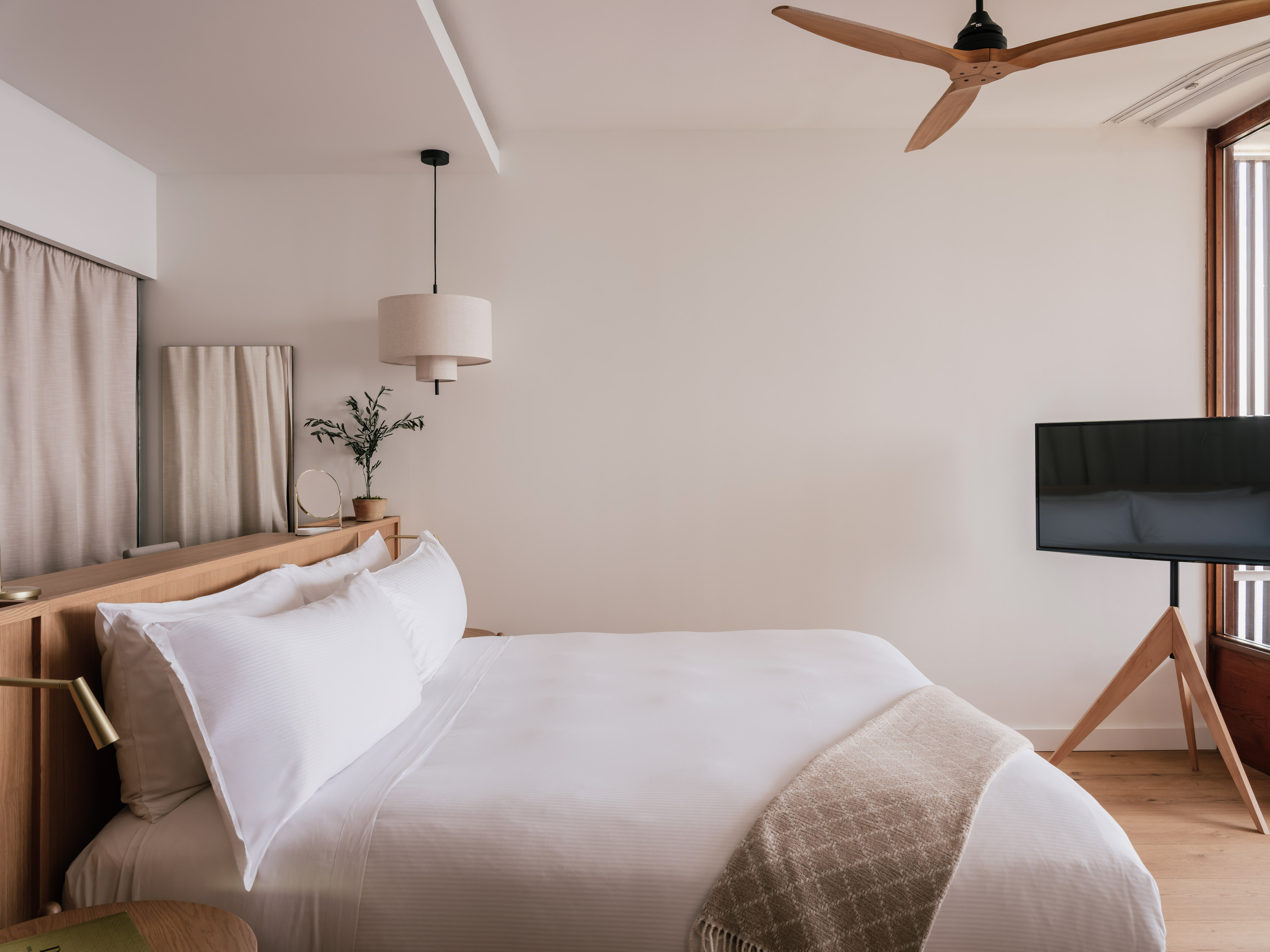 Modern bedroom with white bed, wood headboard, ceiling fan, and TV.