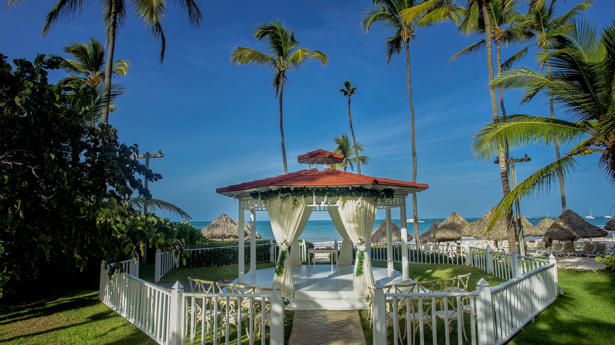 a white gazebo with white curtains and a white fence surrounded by palm trees