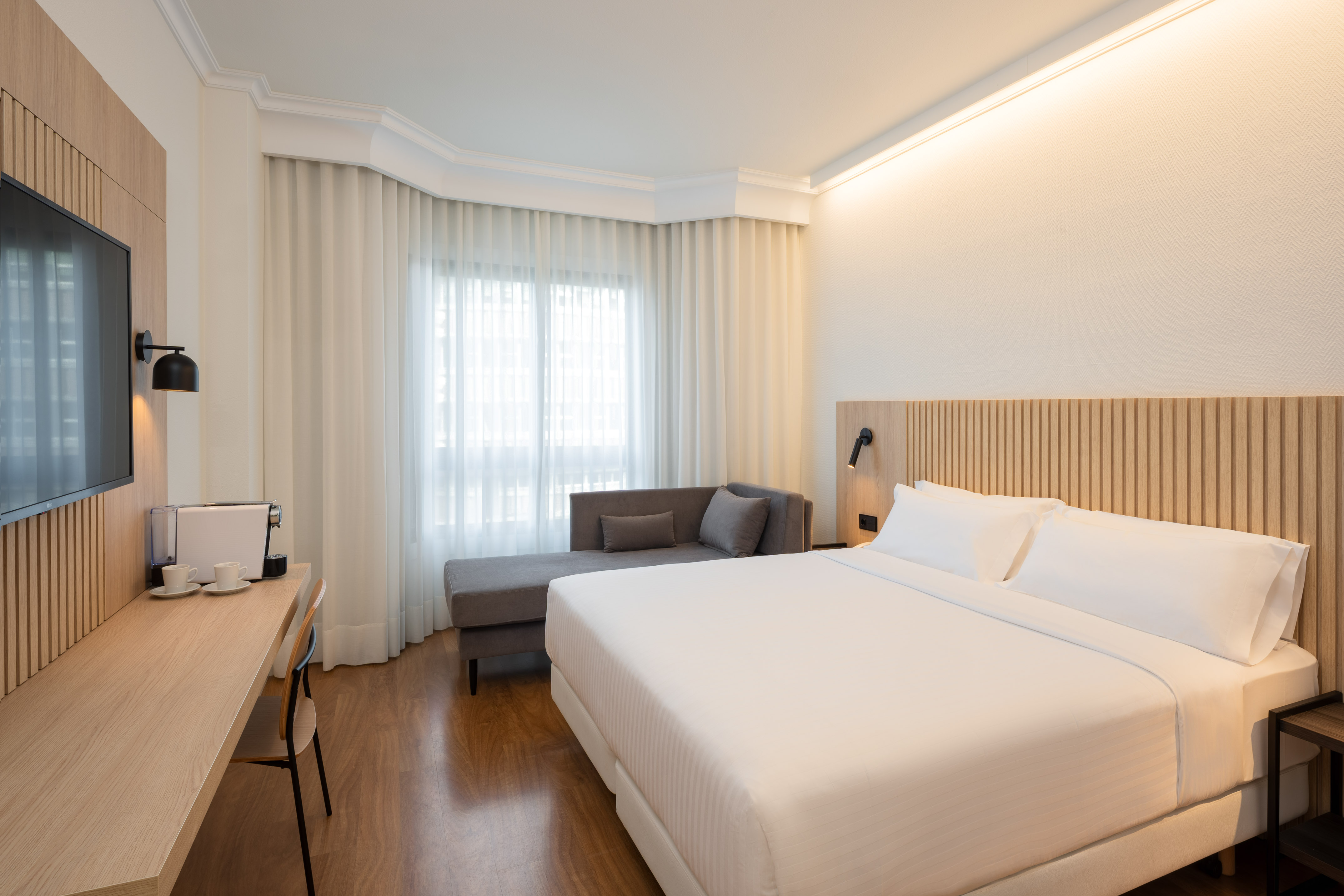 Modern hotel bedroom with white king bed, wooden desk, TV, and grey chaise.