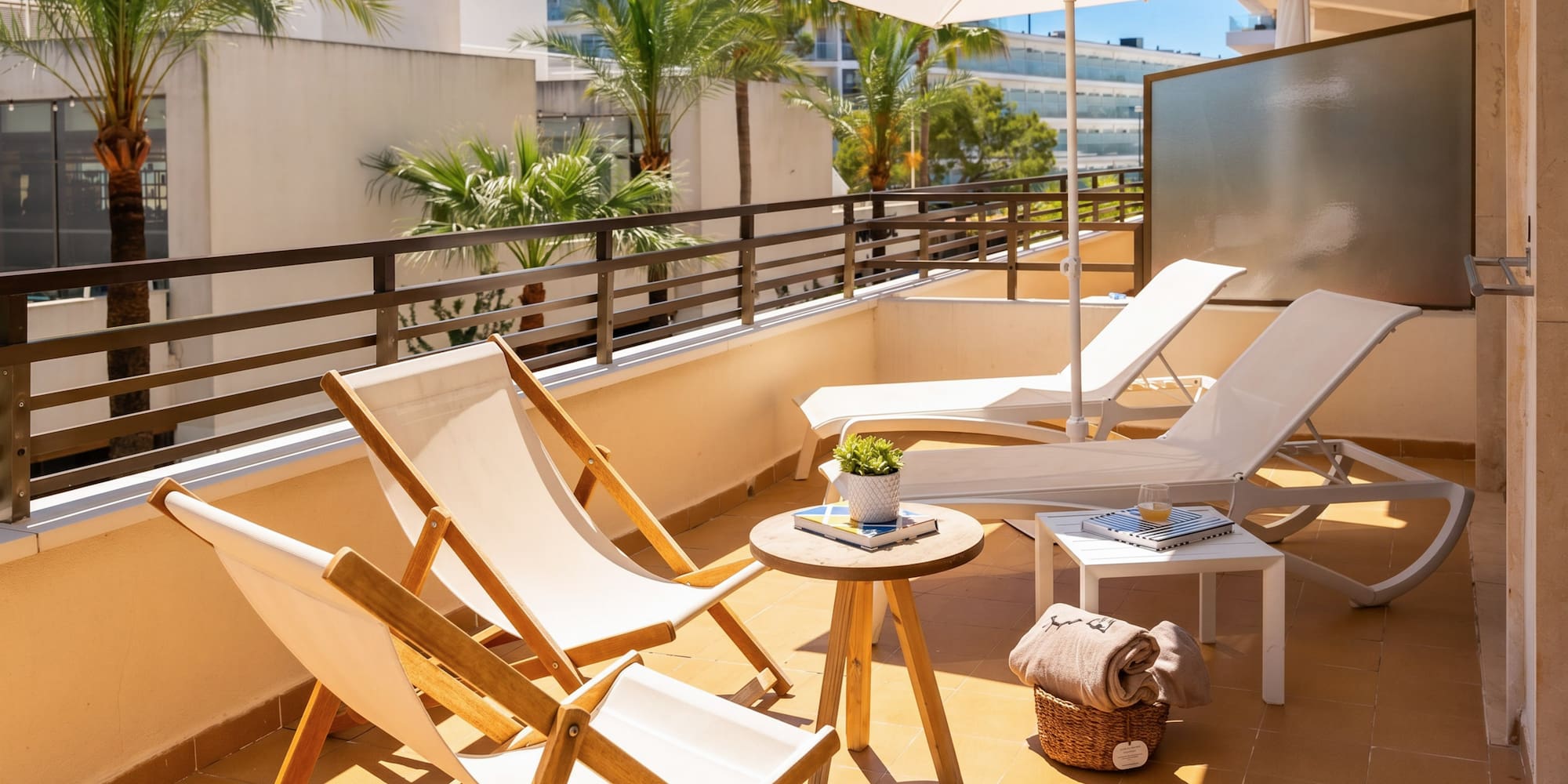 Sunny hotel balcony with white sun loungers, umbrella, and palm trees.